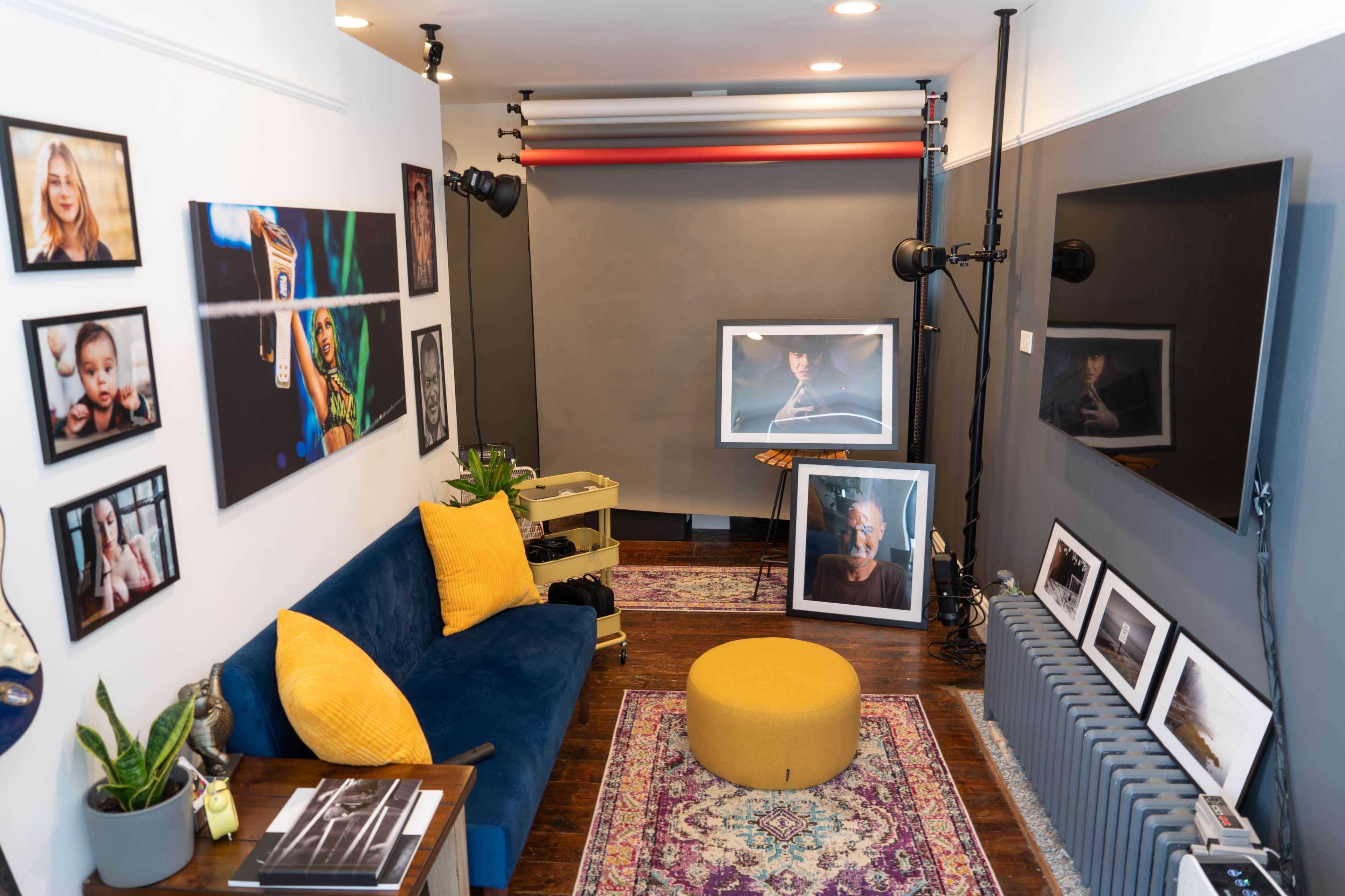 A photography studio with a blue sofa, a yellow pouf, framed photographs on the walls, and various lighting equipment set up.