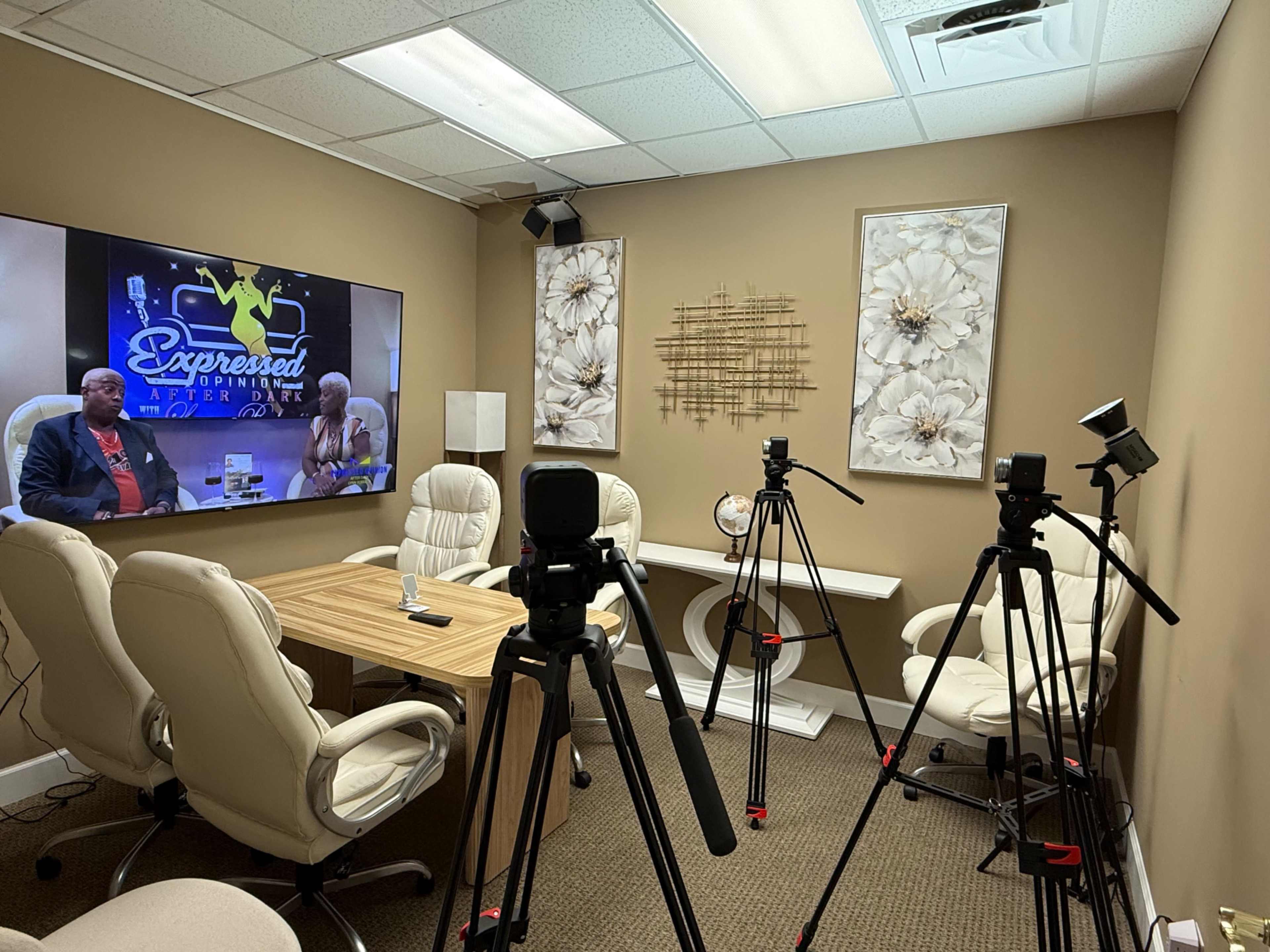 A recording studio is set up with a large screen displaying a show, two chairs facing each other, and several cameras on tripods aimed at the seating area.