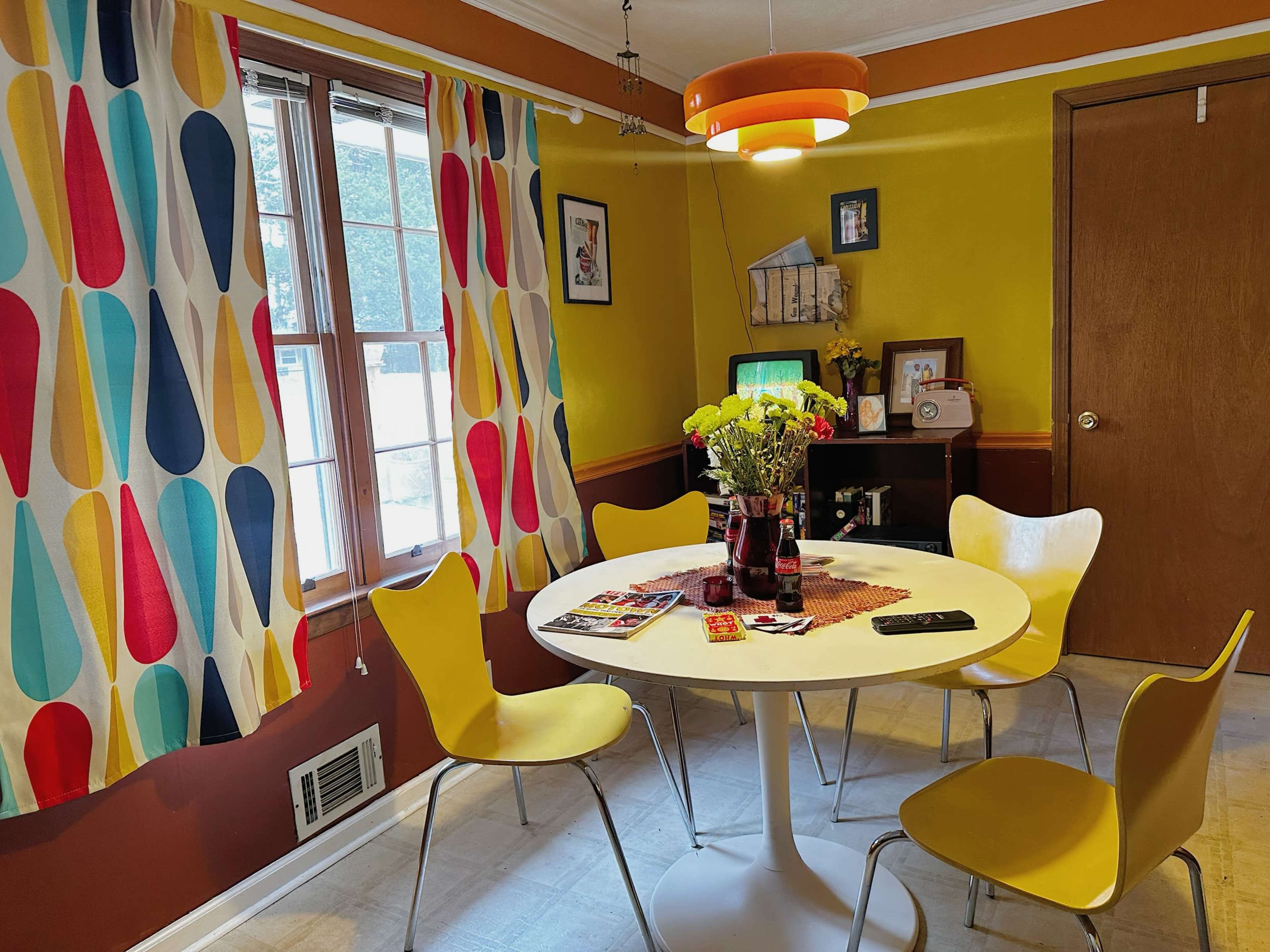 A round white table surrounded by four yellow chairs is positioned in a brightly decorated dining area with colorful curtains and a vintage TV.