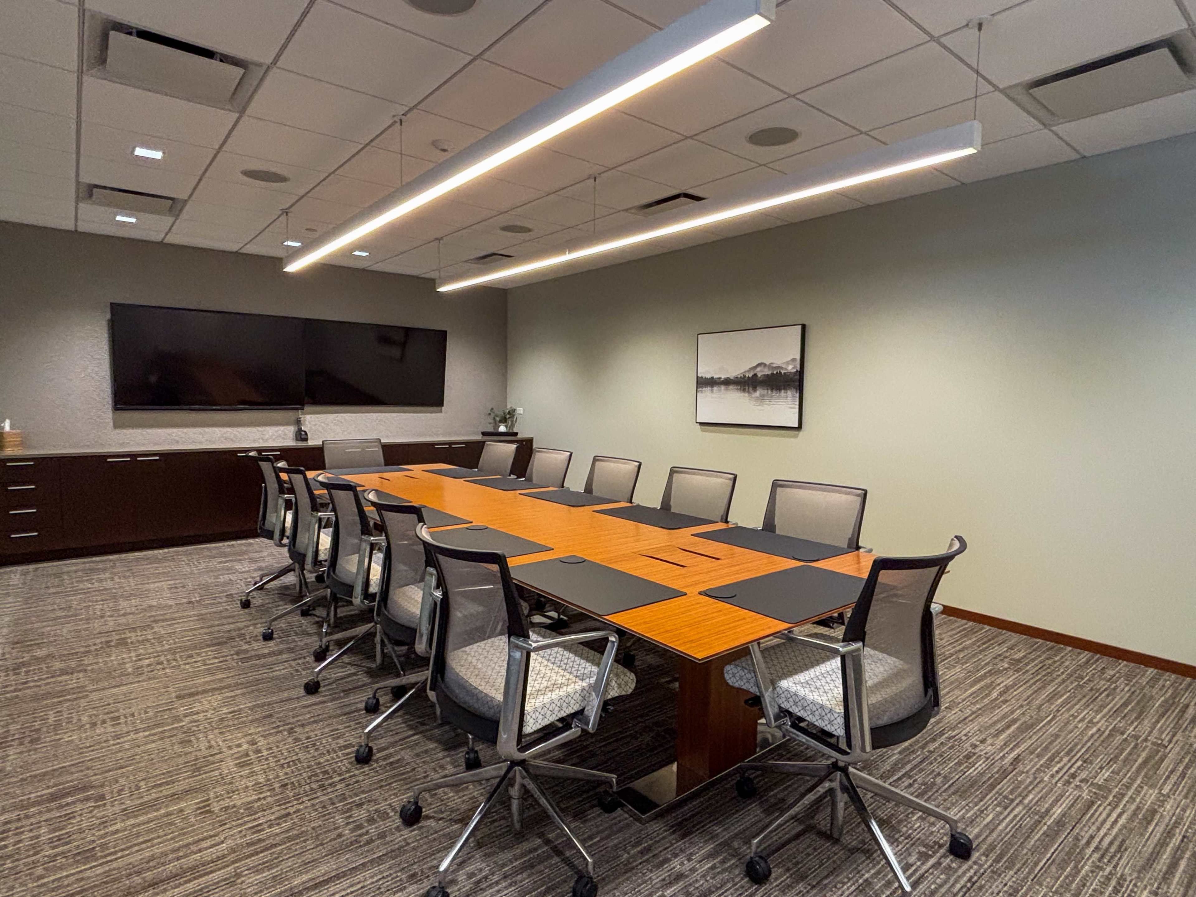 The image shows a modern conference room equipped with a long wooden table surrounded by ergonomic chairs, a large screen on the wall, and ambient lighting.