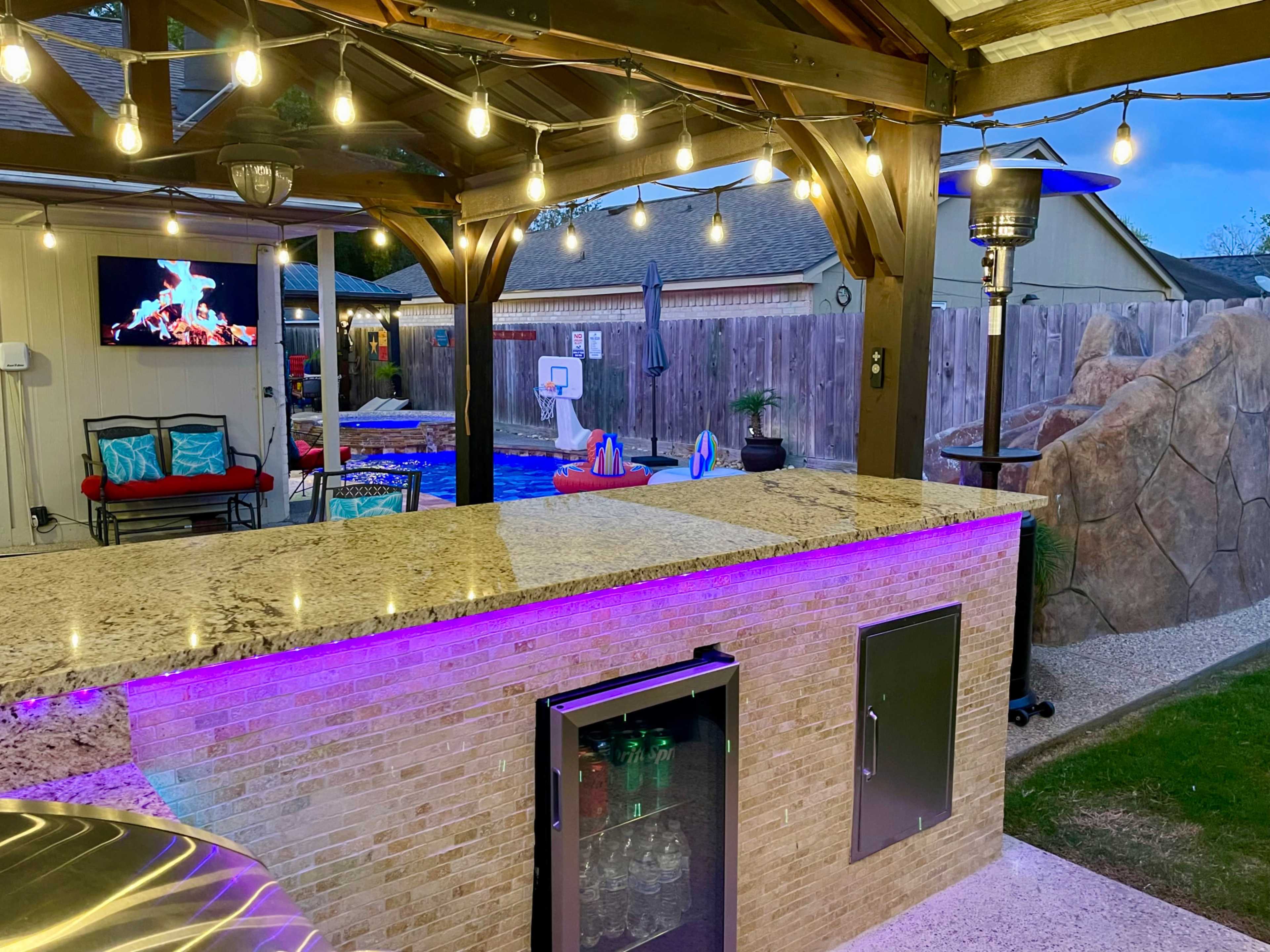 An outdoor entertainment area with a granite bar, a beverage fridge, a large screen, and illuminated string lights, overlooking a swimming pool with a basketball hoop.