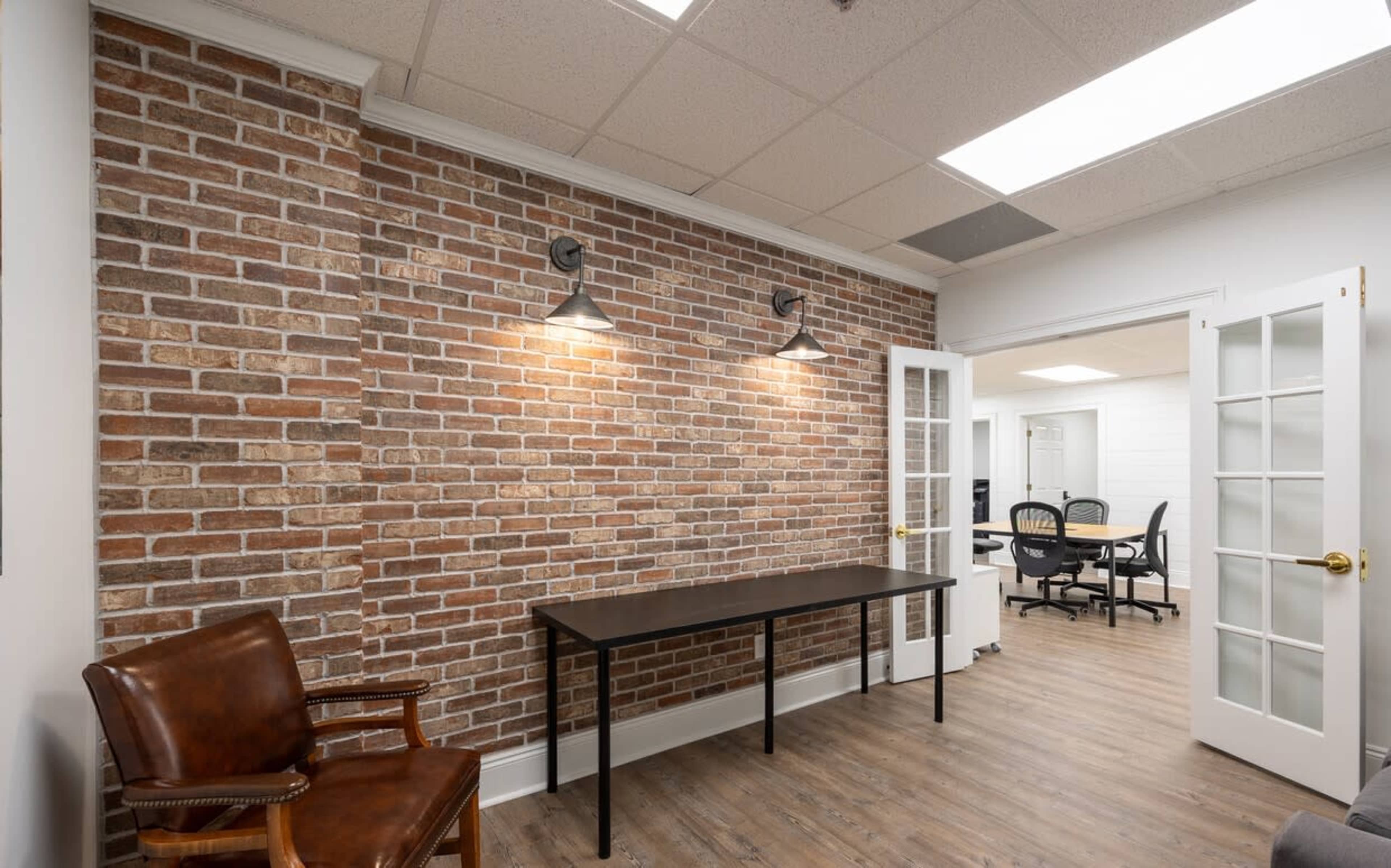 A small room with exposed brick walls, a wooden table, a brown chair, and a door leading to another office space.
