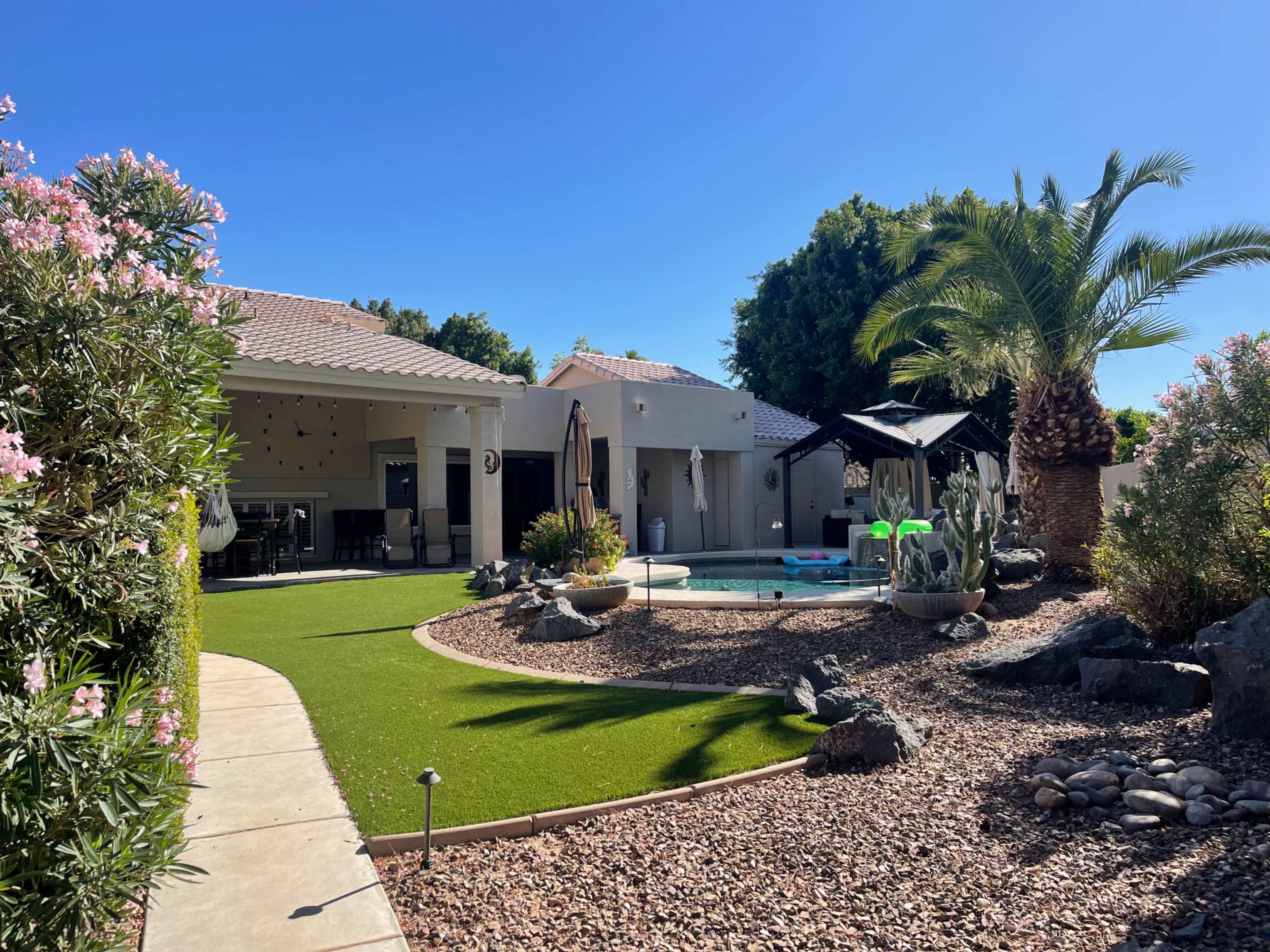 A landscaped backyard with a pool, palm trees, and a gazebo surrounded by gravel and ornamental plants.