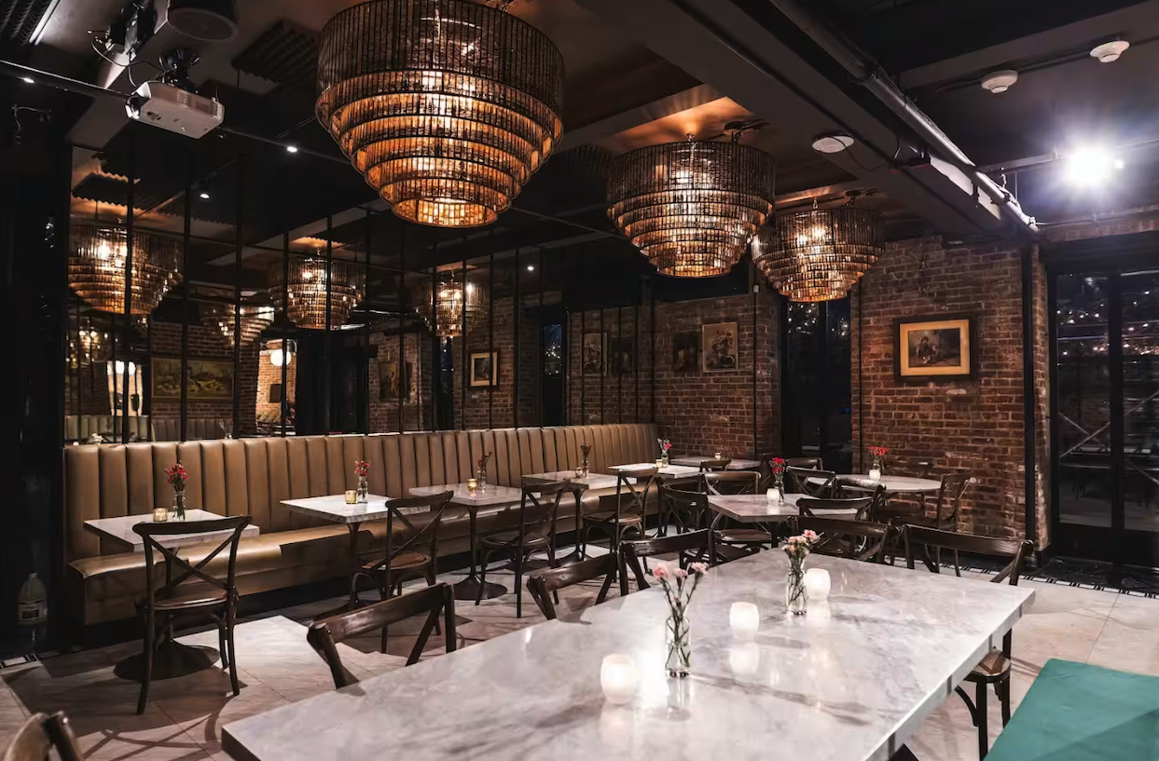 The image shows a modern restaurant interior featuring marble-top tables, wooden chairs, decorative chandeliers, and a brick wall backdrop.