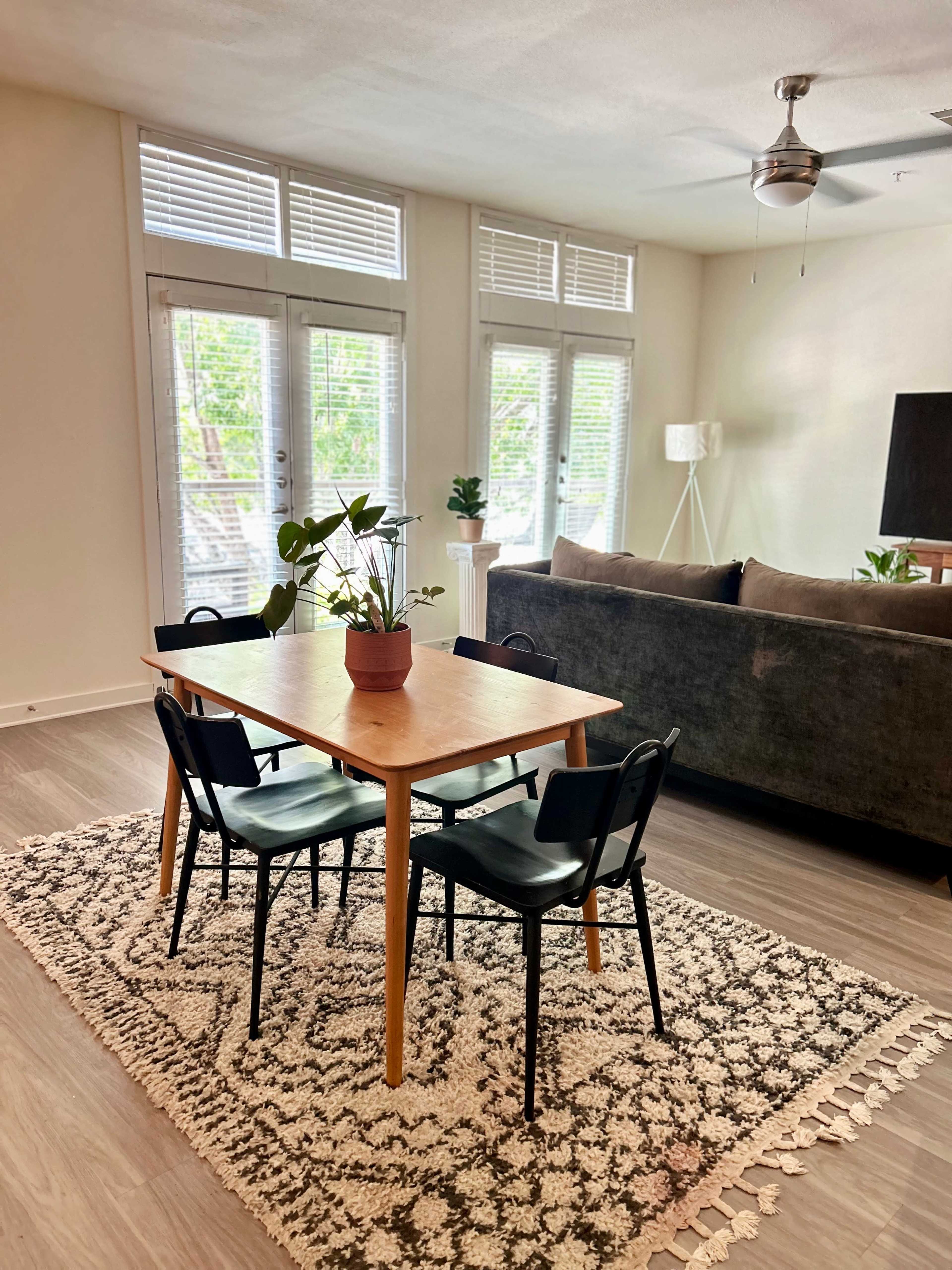 A wooden dining table with four black chairs sits on a patterned rug in a bright living space featuring large windows and a gray couch.
