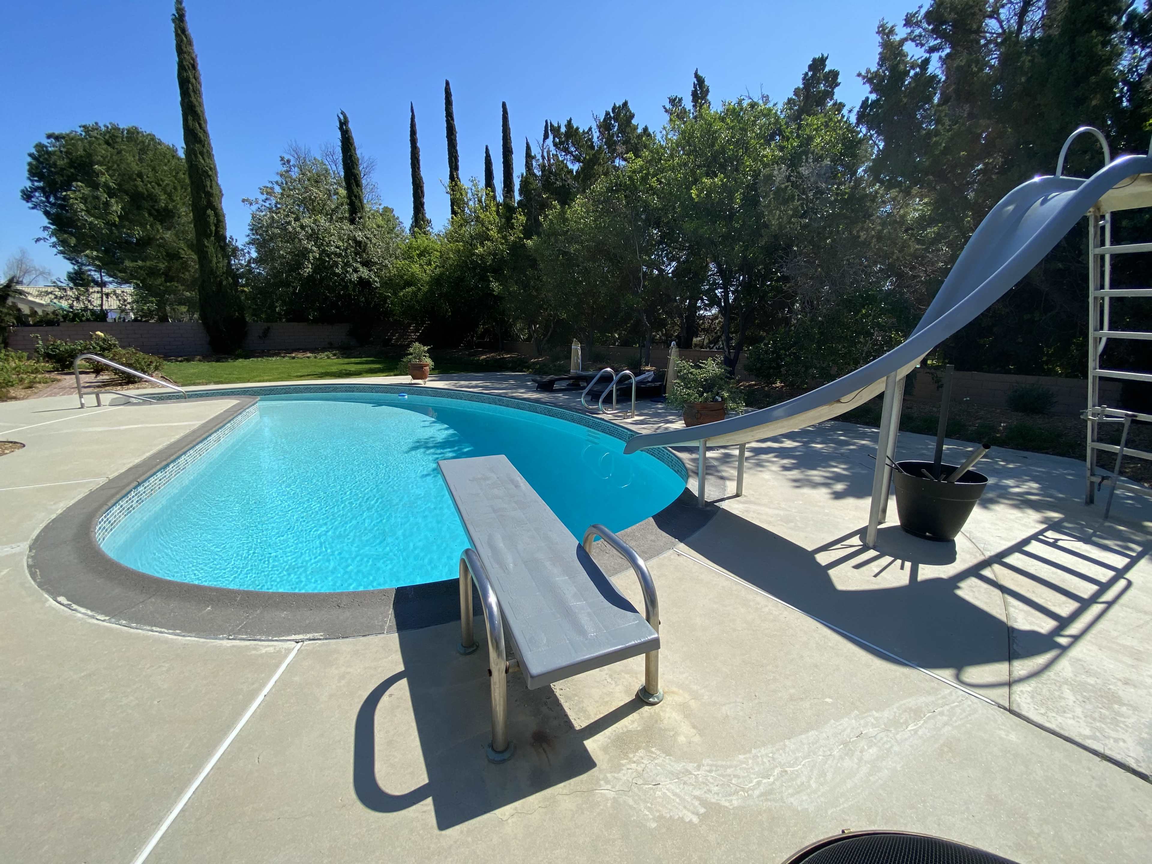 A swimming pool with a slide, a diving board, and tall trees surrounding the area.
