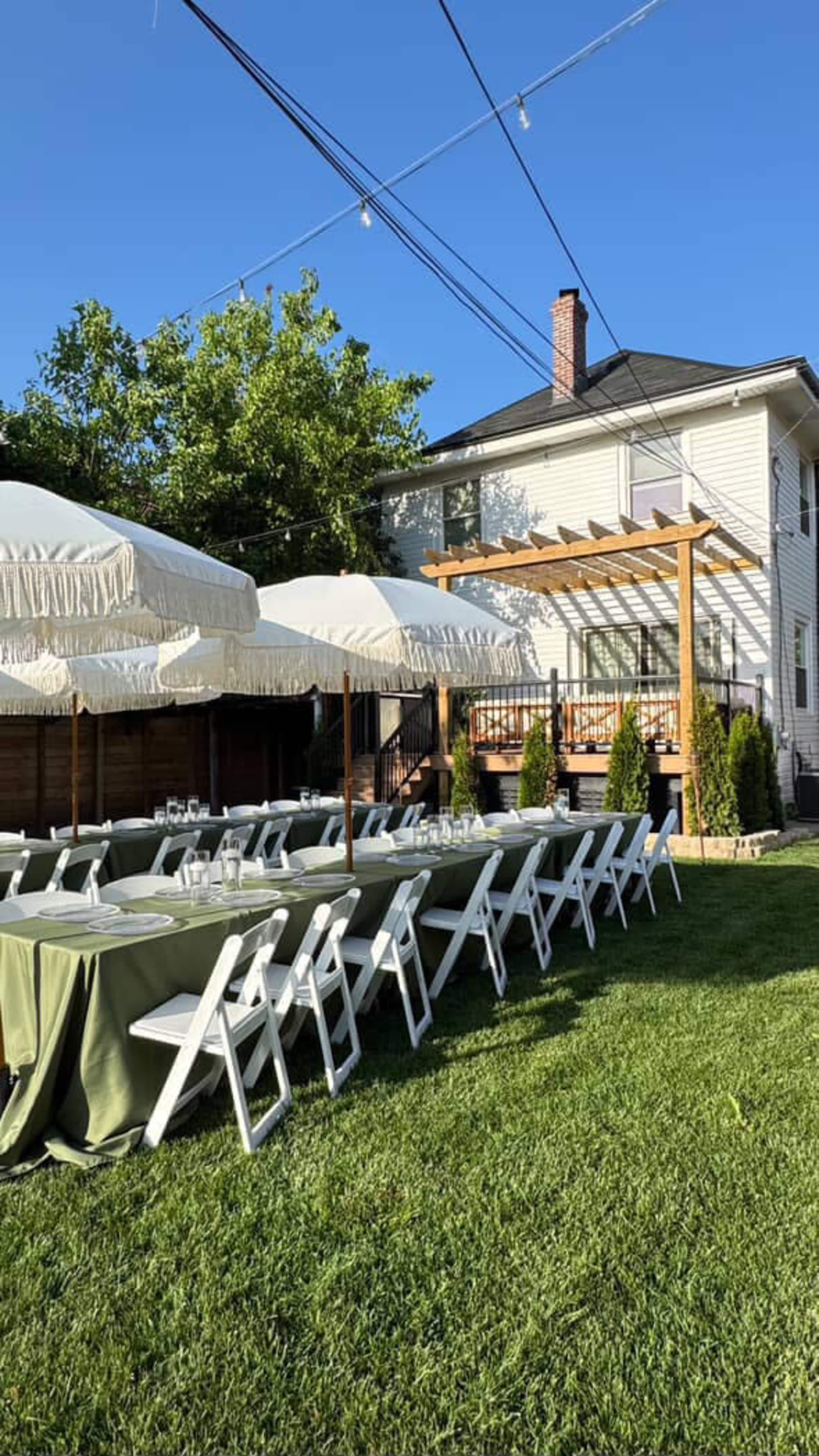 A set of long tables with green tablecloths and white folding chairs is arranged under large white umbrellas on a grassy area near a house with a deck.