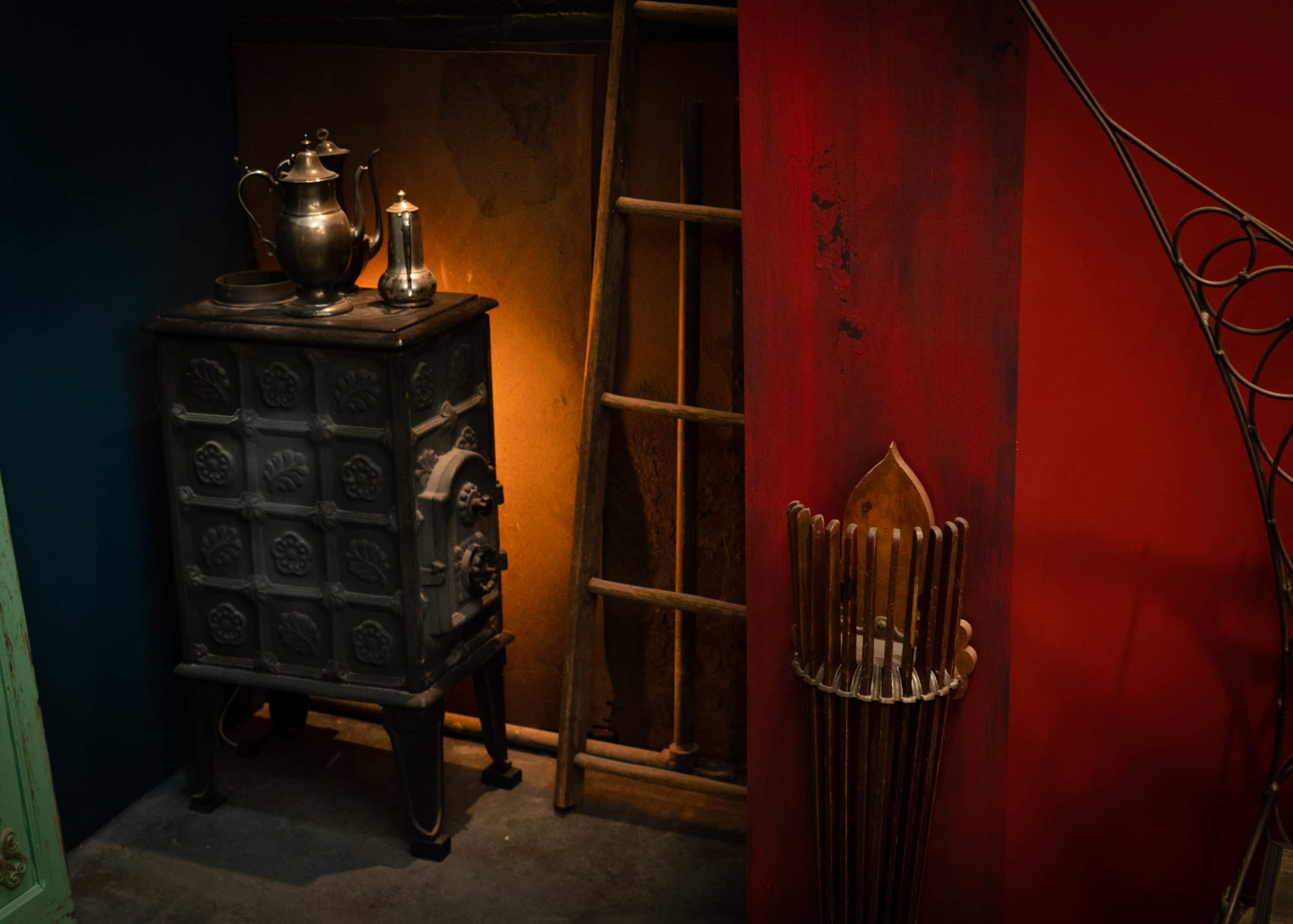 The image depicts a rustic setting featuring a cast-iron stove, a metallic teapot, a wooden ladder, and a decorative wall with a striking color contrast.