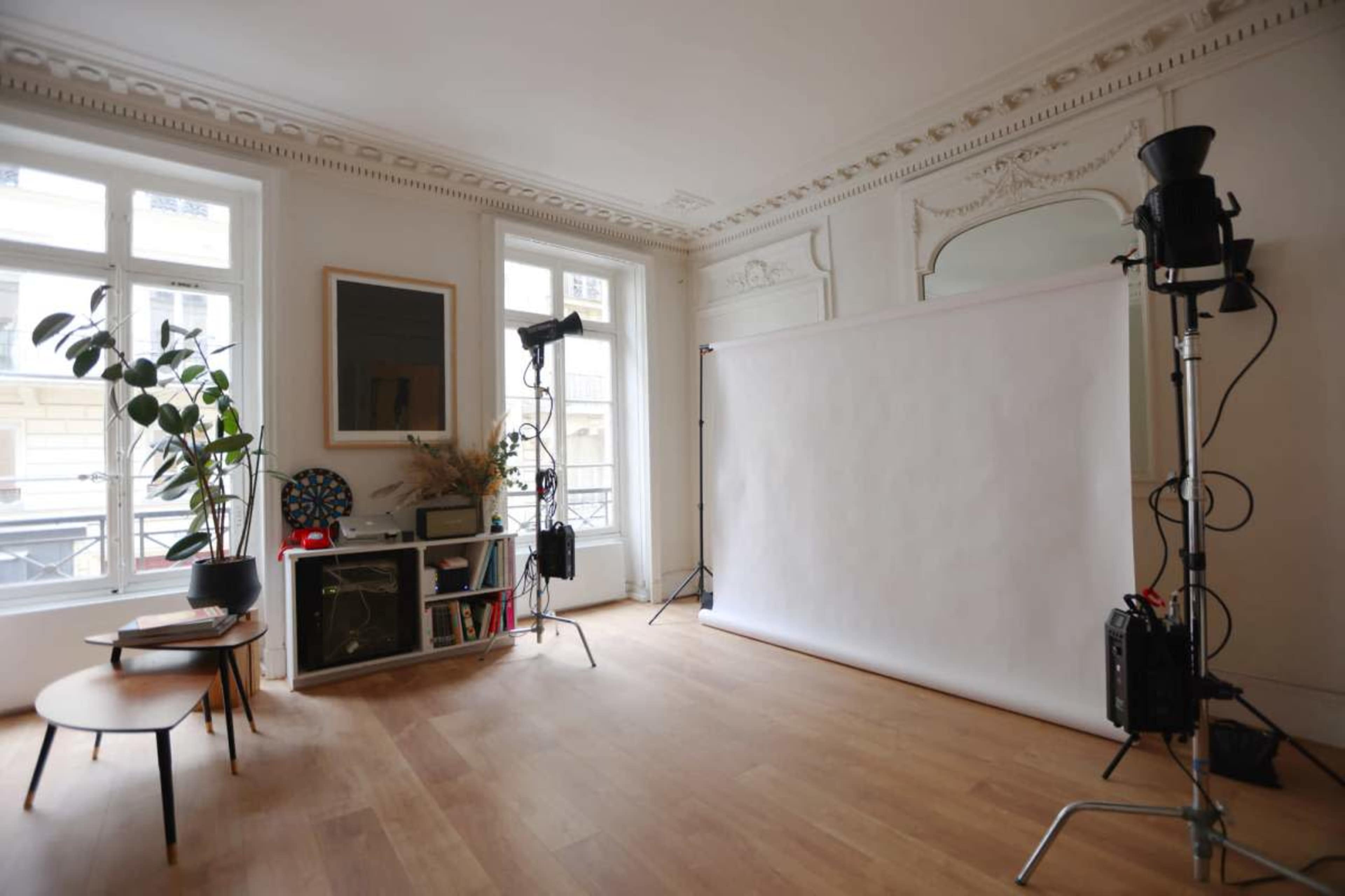 The image shows a bright, airy studio space with a white backdrop, photography lights, a plant, and minimalist furniture.