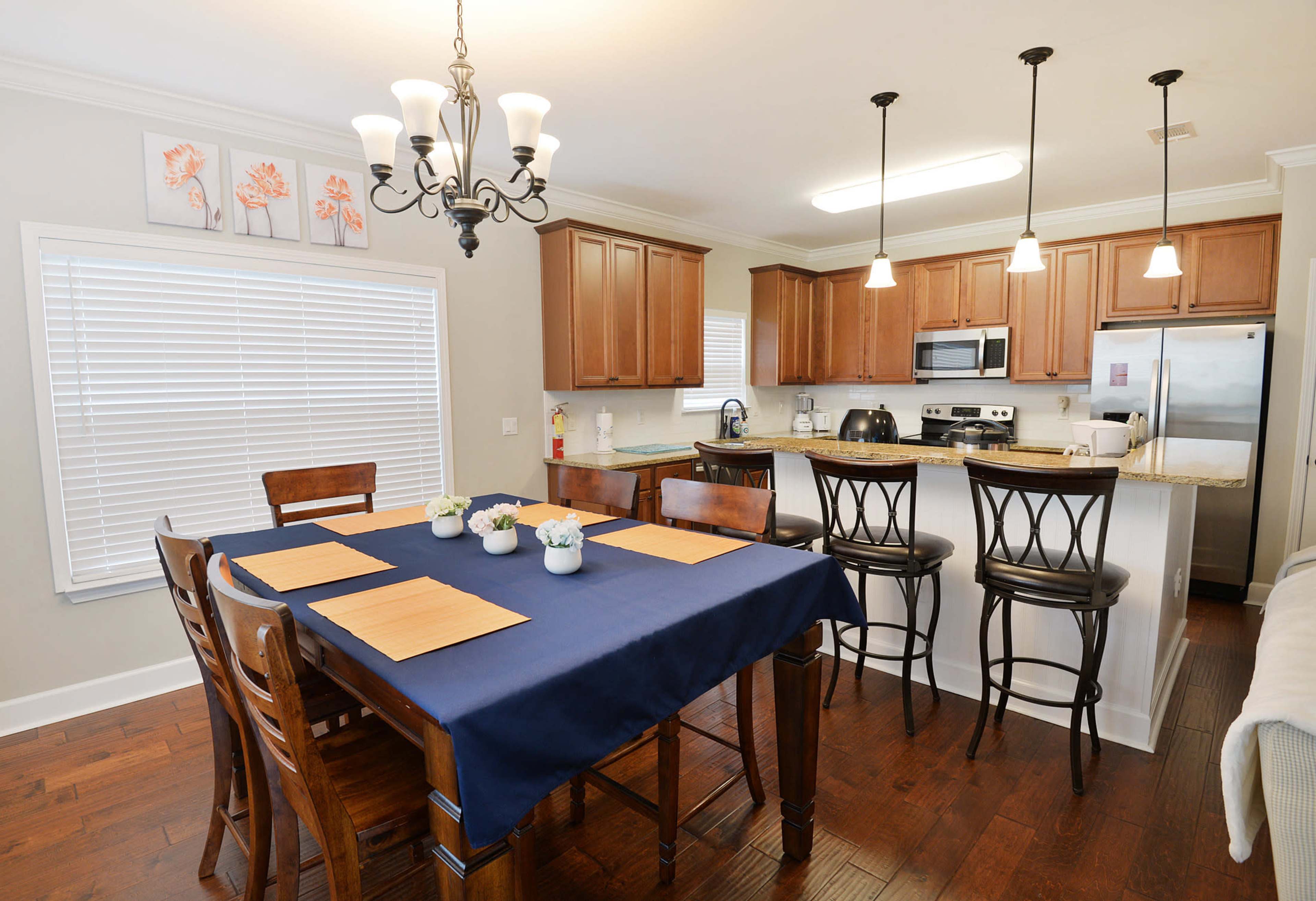 A kitchen and dining area with a dark wooden table set for a meal, featuring blue placemats and surrounded by wooden chairs, adjacent to a modern kitchen with wooden cabinetry and stainless steel appliances.