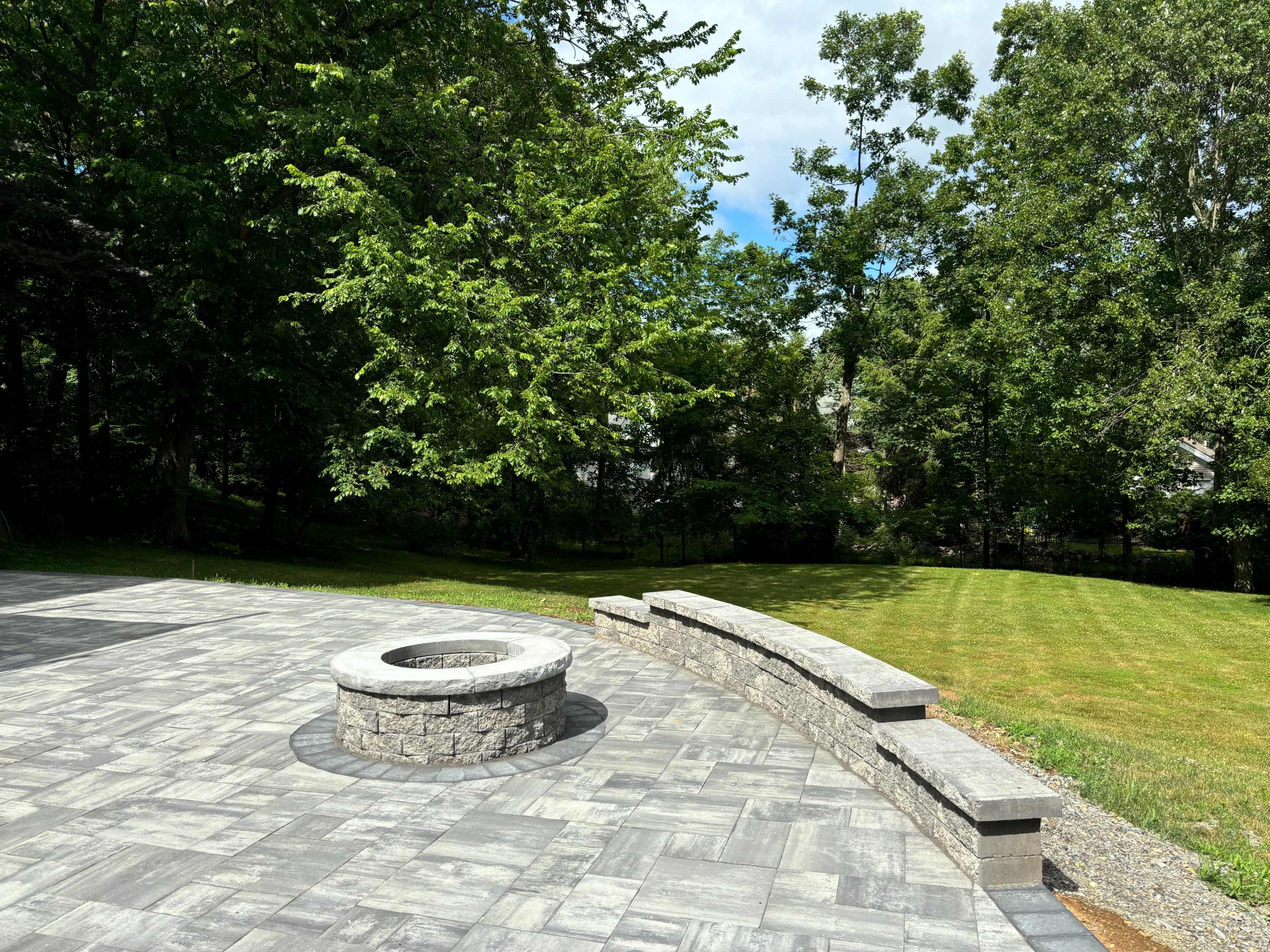 A stone fire pit surrounded by a paved patio and a curved stone bench, set against a backdrop of green trees and grass.