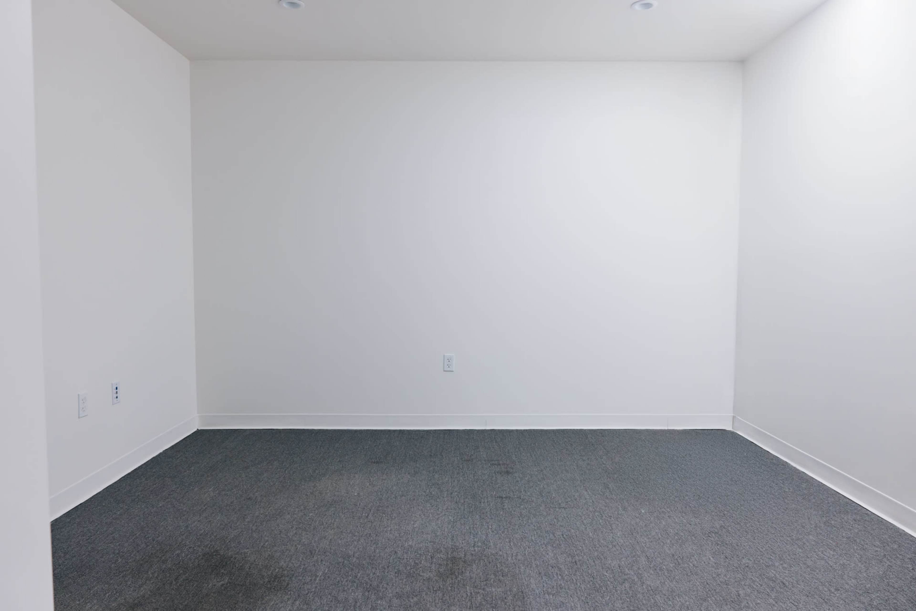 The image shows a sparsely furnished room with gray carpet and white walls, featuring minimal decor and empty space.