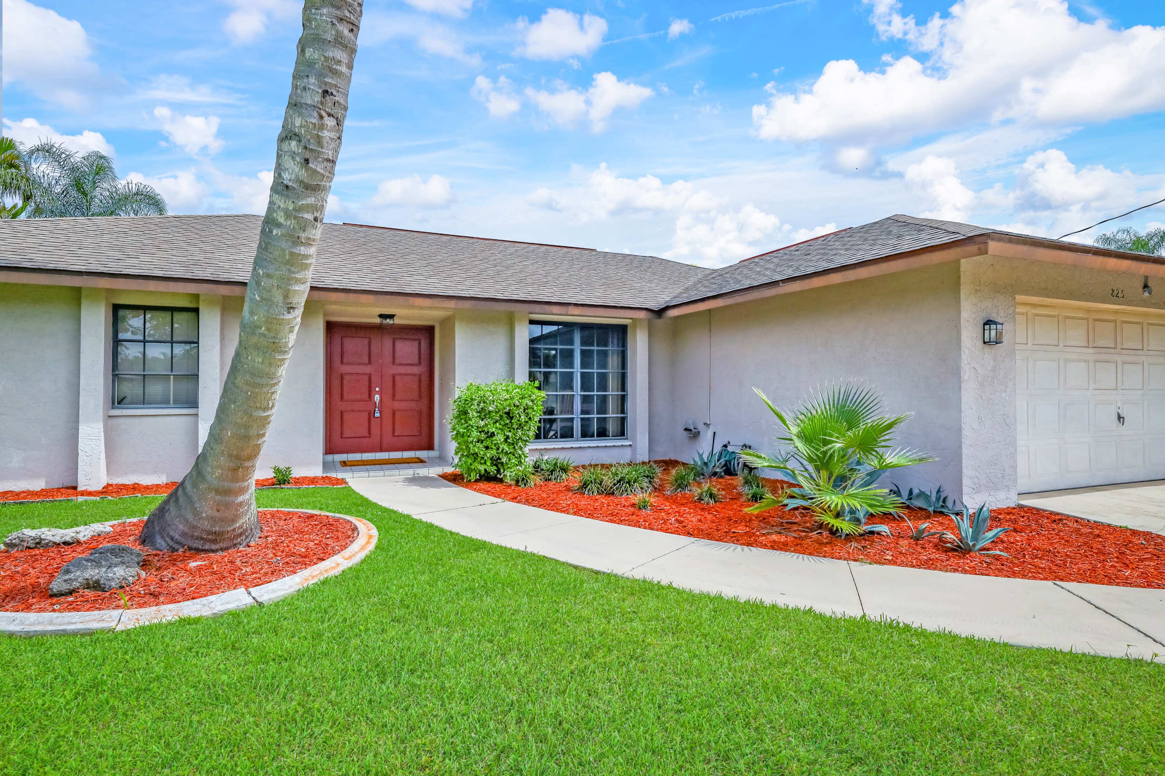 A single-story house with a red front door, surrounded by a well-maintained lawn and decorative landscaping featuring red mulch and tropical plants.