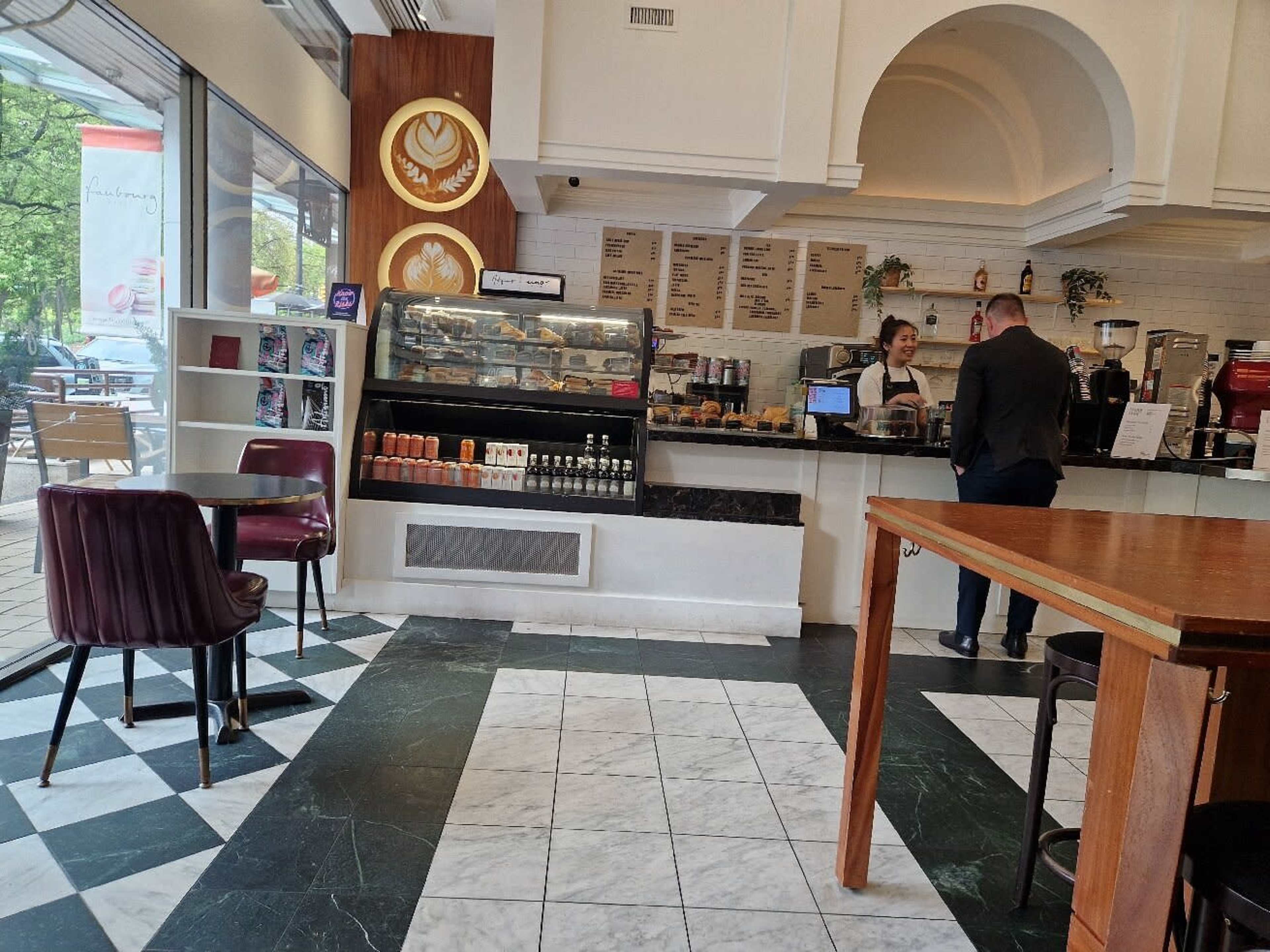 A coffee shop interior features a counter with pastries and beverages on display, while a customer interacts with the barista.