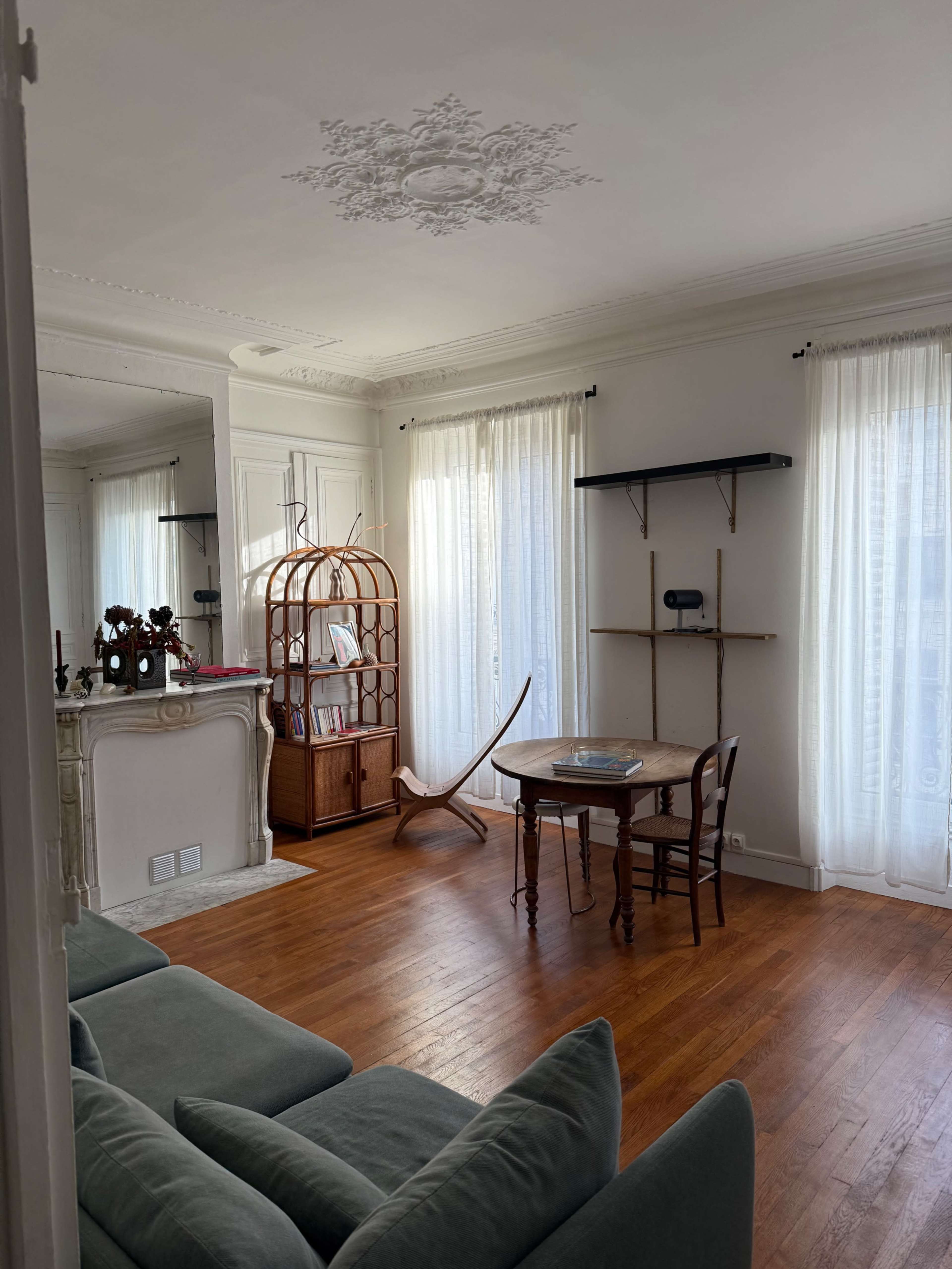 The image shows a cozy living room with wooden floors, a small round table, a comfortable sofa, and a decorative wicker shelving unit against the wall.