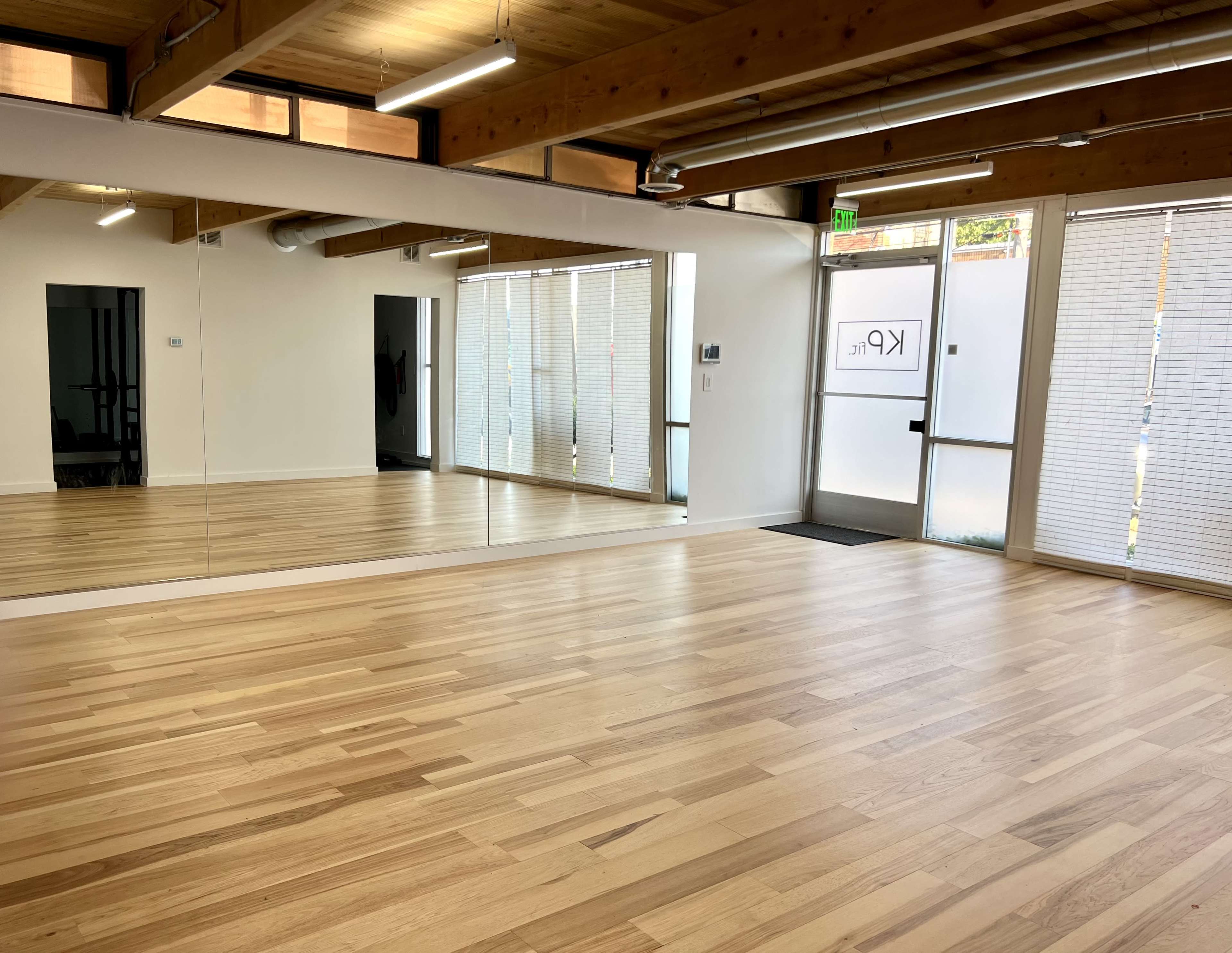 A spacious, empty studio with wooden flooring and a large mirror along one wall, featuring natural light from windows and a glass entrance door.