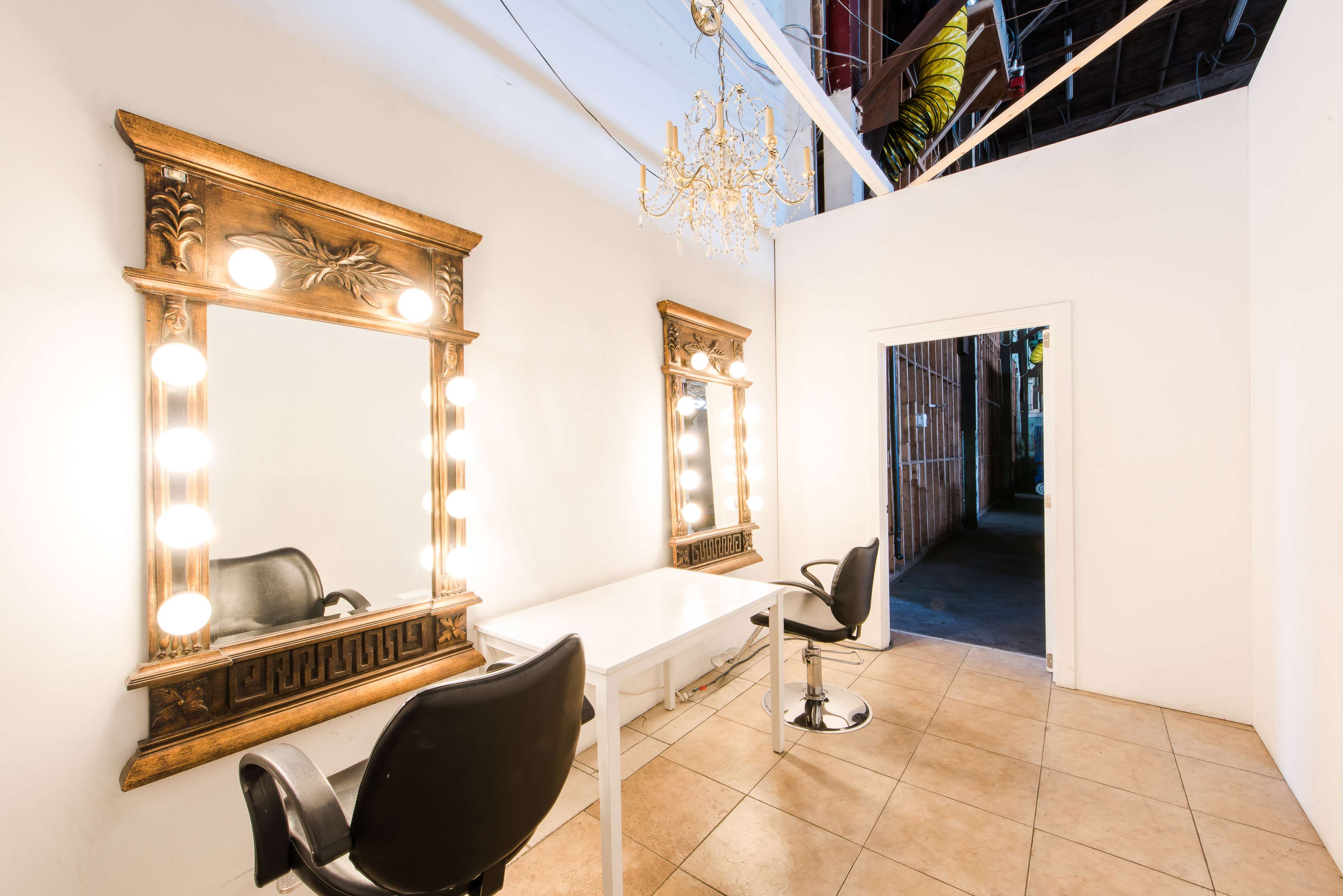 The image shows a well-lit room featuring two large mirrors with lights, a table, and two black chairs, with an open doorway leading to an adjacent space.