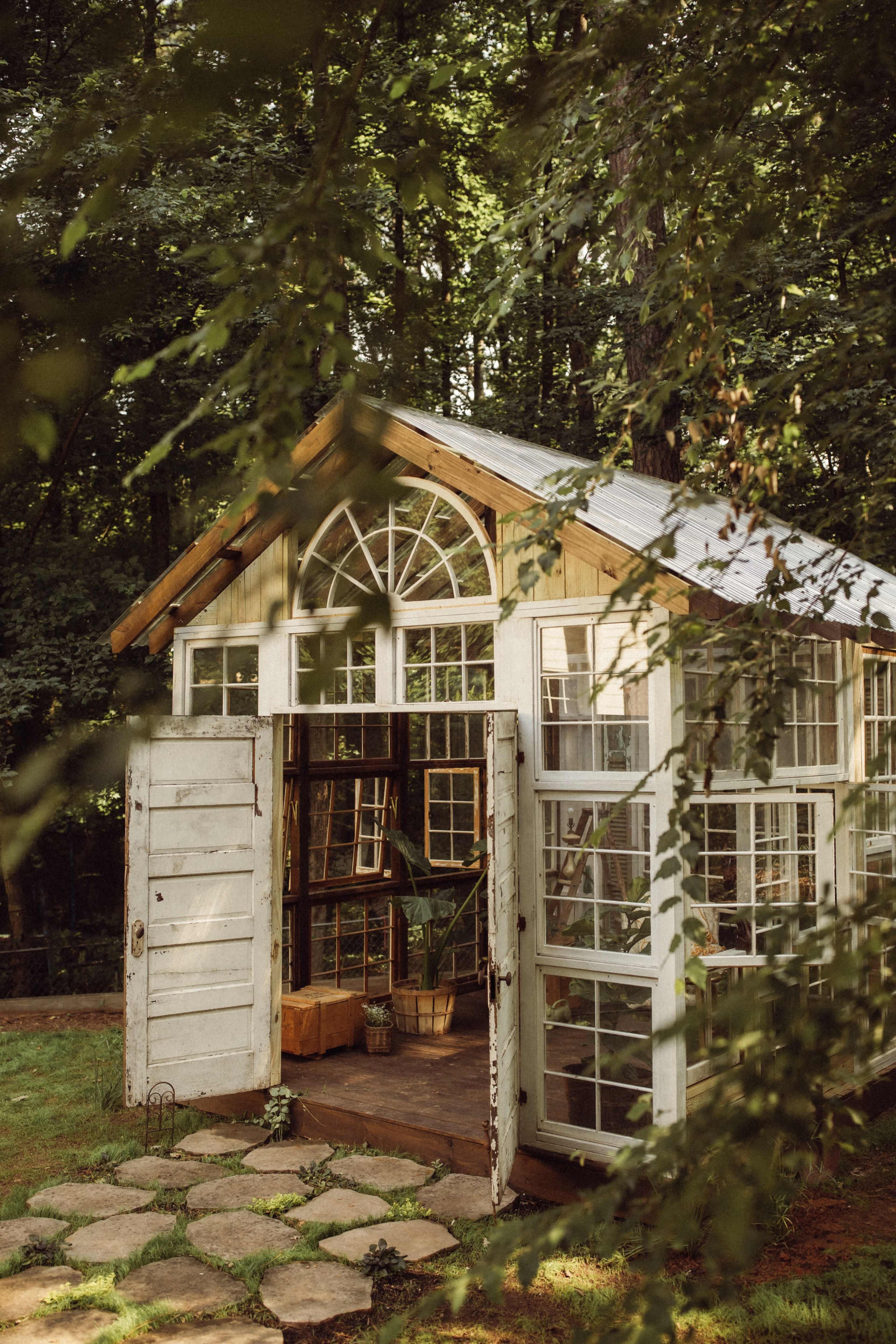 A small greenhouse made of wood and glass stands in a wooded area, featuring double doors open to a stone pathway and surrounded by greenery.