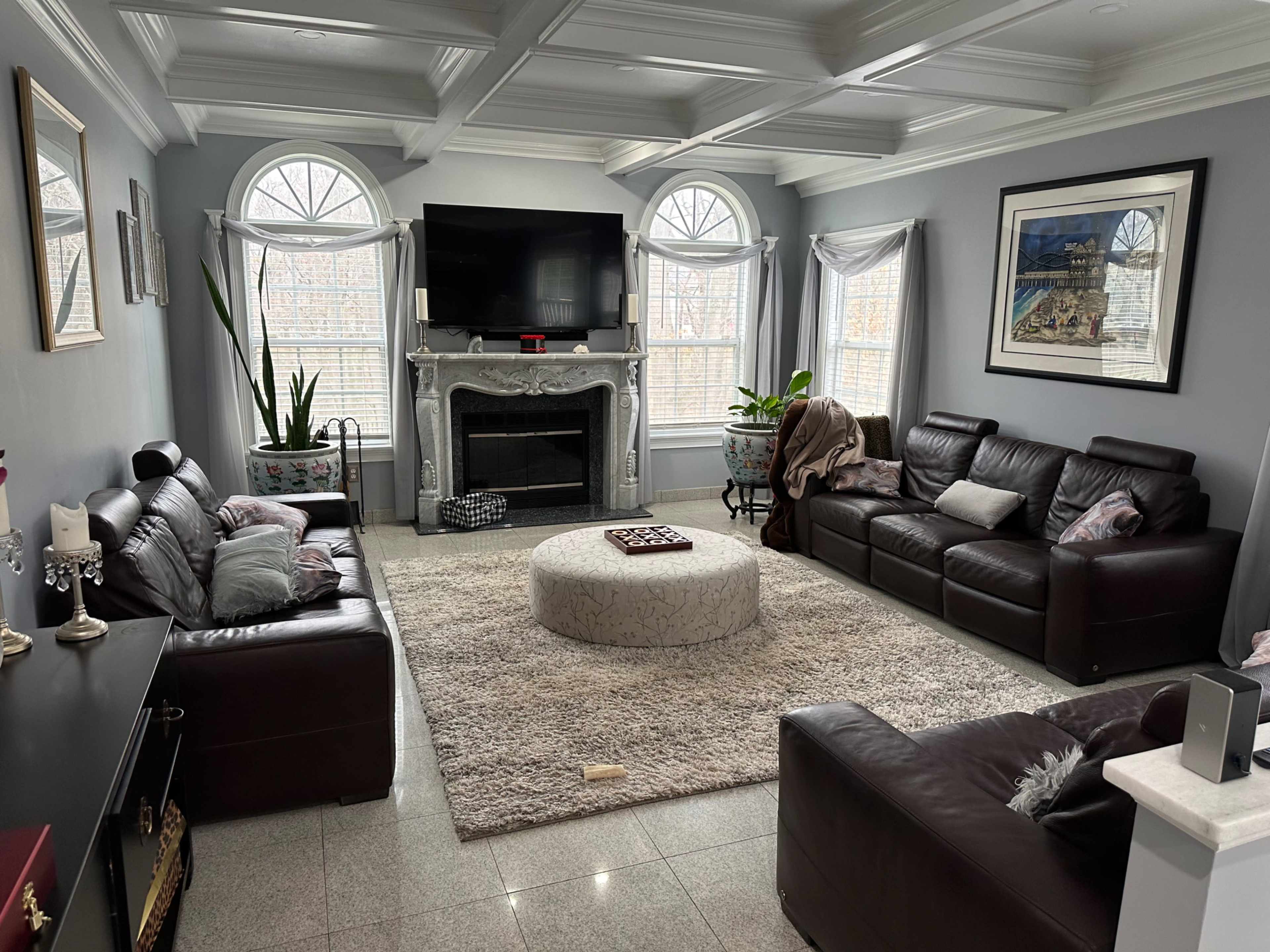 The living room features two dark leather sofas facing a central circular coffee table, with a large television mounted above a decorative fireplace and windows allowing natural light.