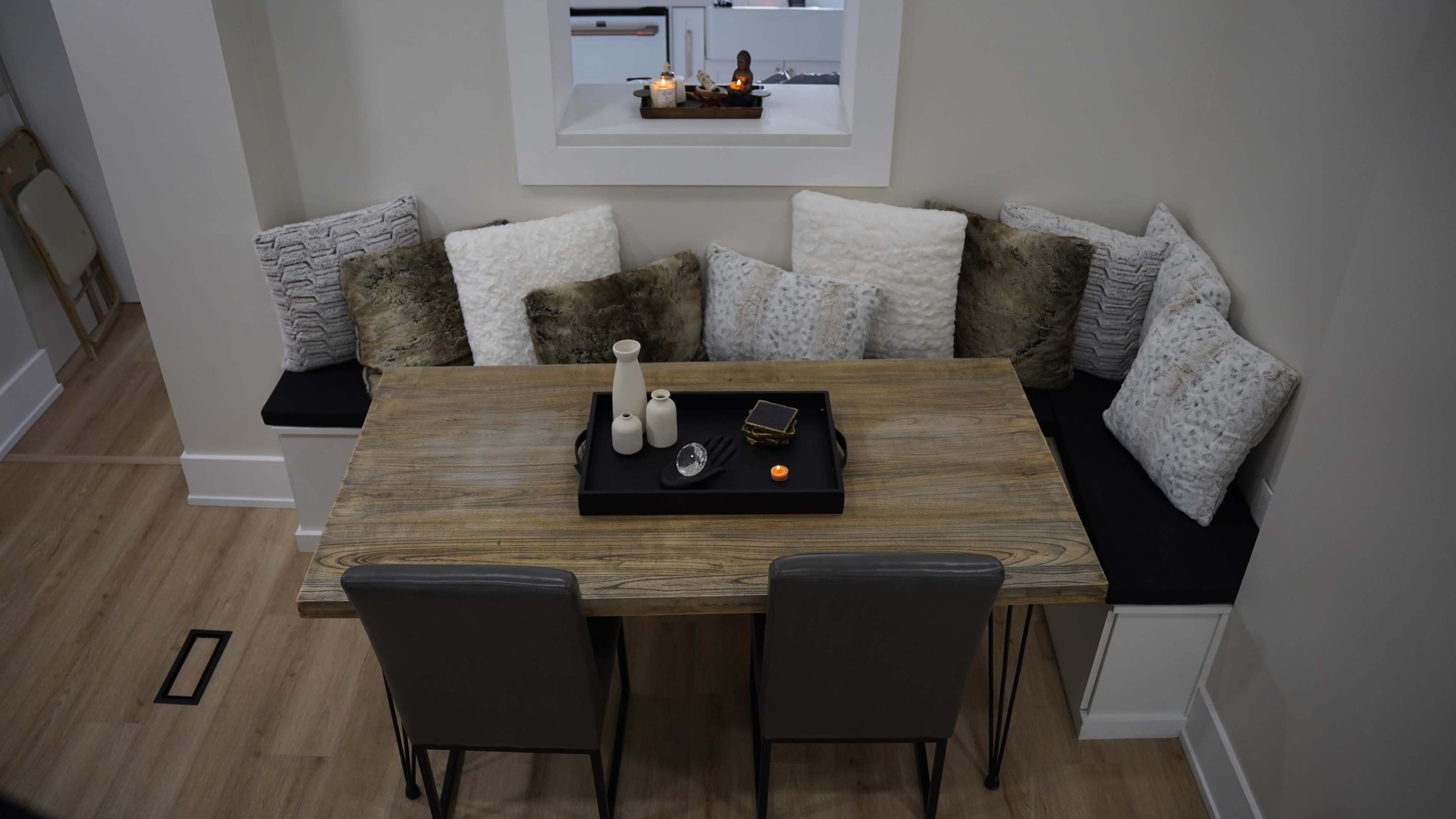 A wooden dining table with a black tray sits in the center of a cozy nook surrounded by plush pillows.