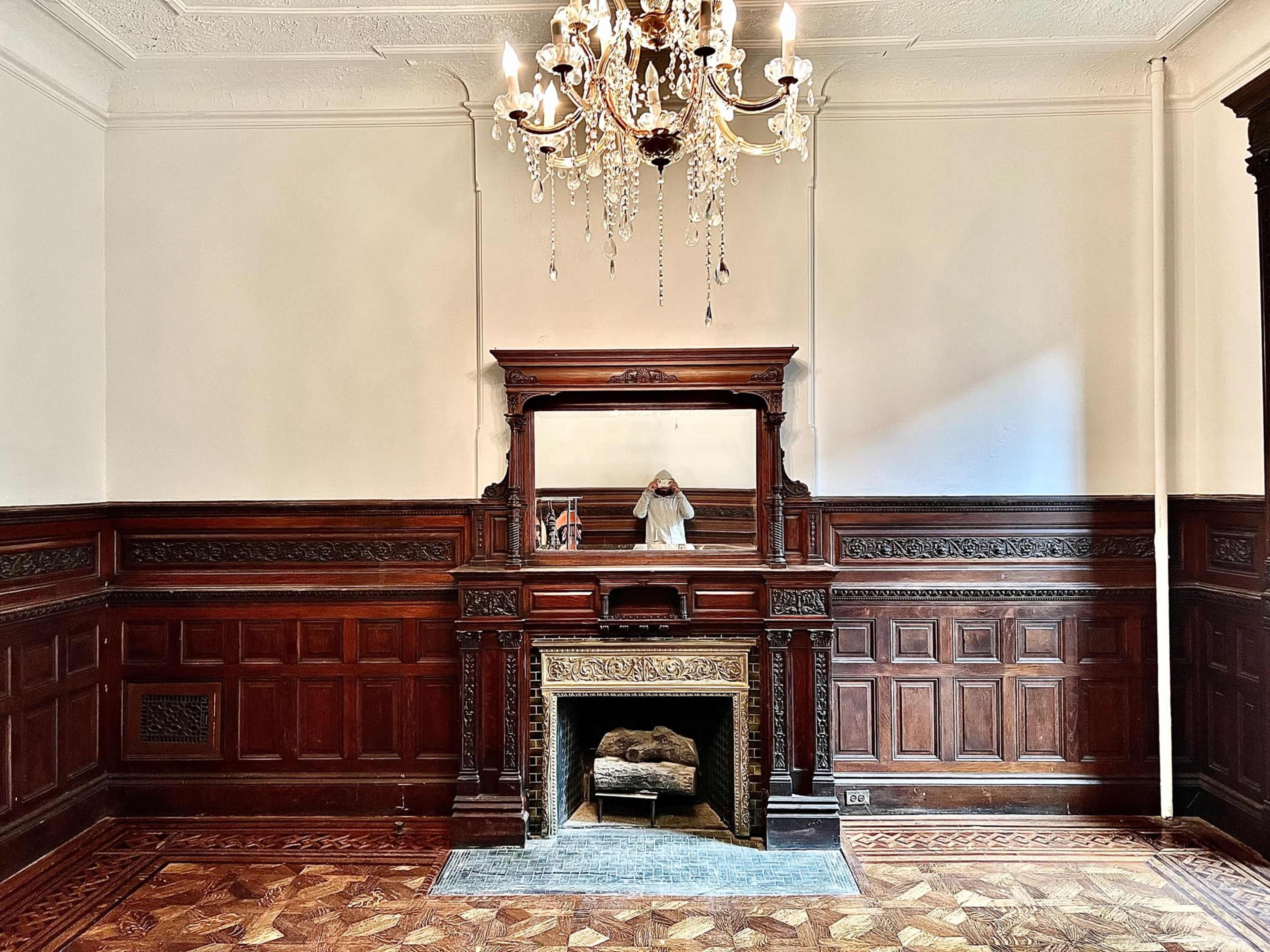 The image shows a vintage room with dark wood paneling, a fireplace with an ornate mantle, and a chandelier hanging from the ceiling.