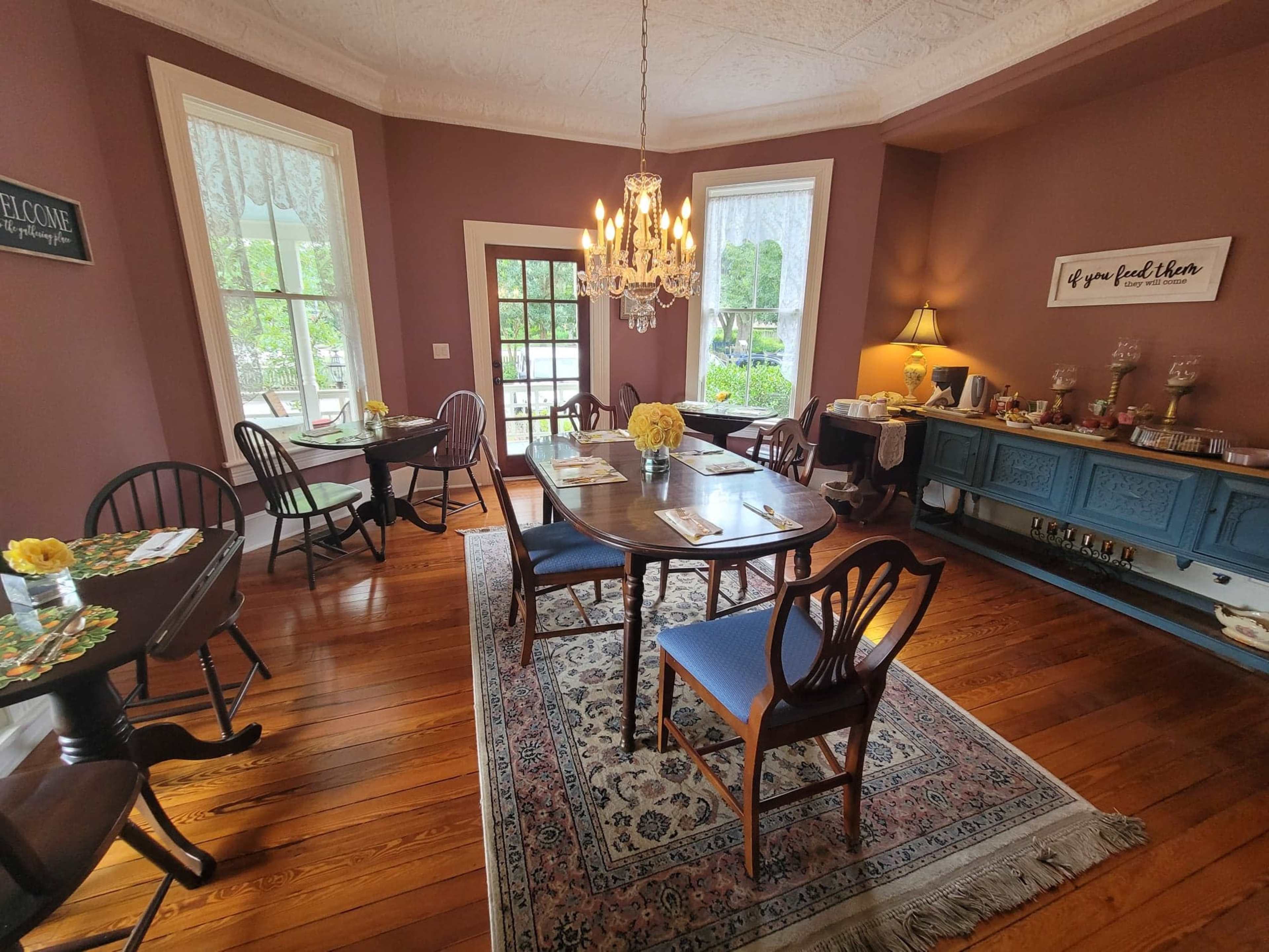 A well-furnished dining room with several tables set for guests, a chandelier hanging from the ceiling, and a sideboard displaying food items.
