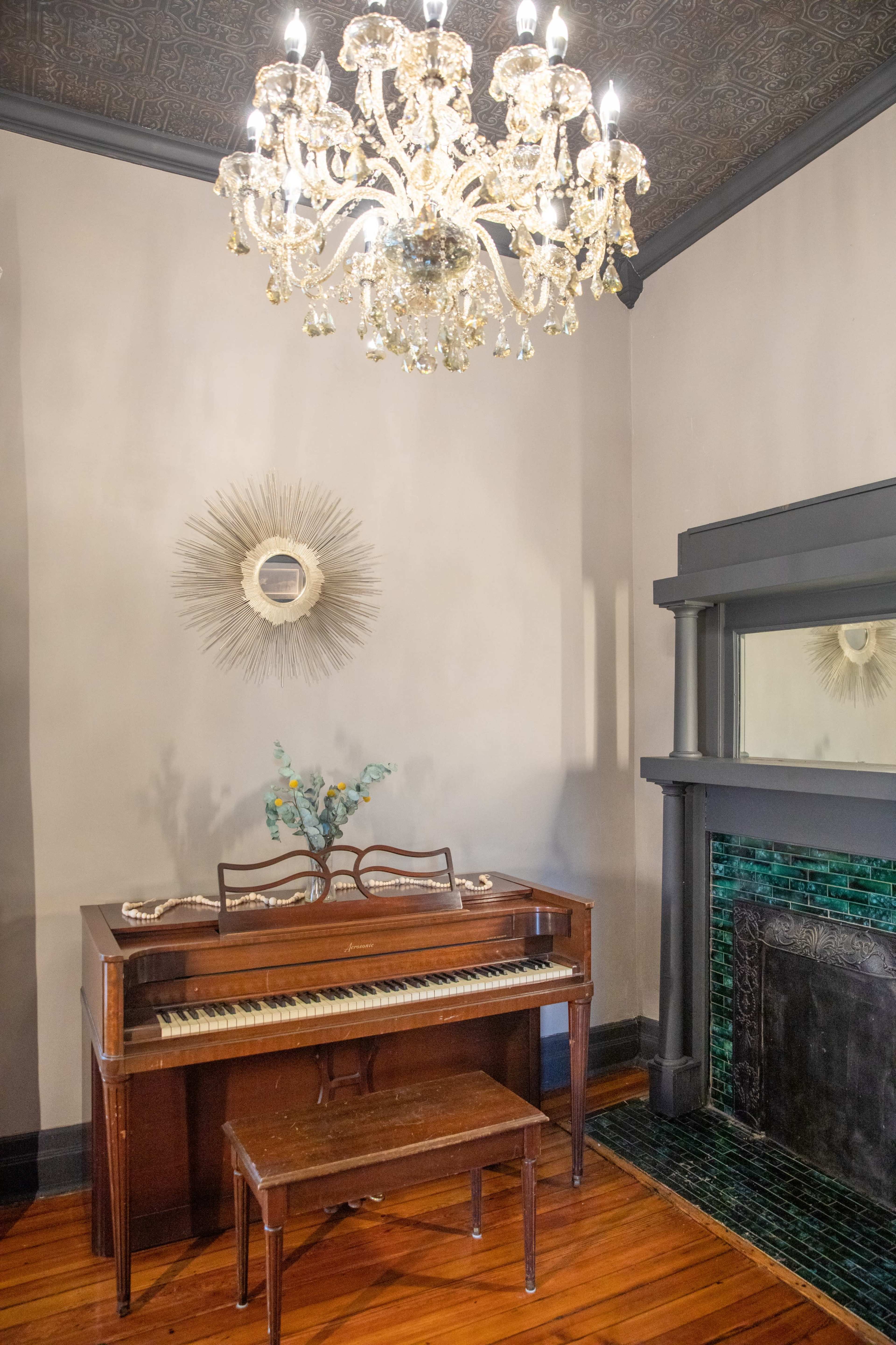 A vintage piano sits beside a decorative fireplace in a room adorned with a chandelier and a circular mirror.