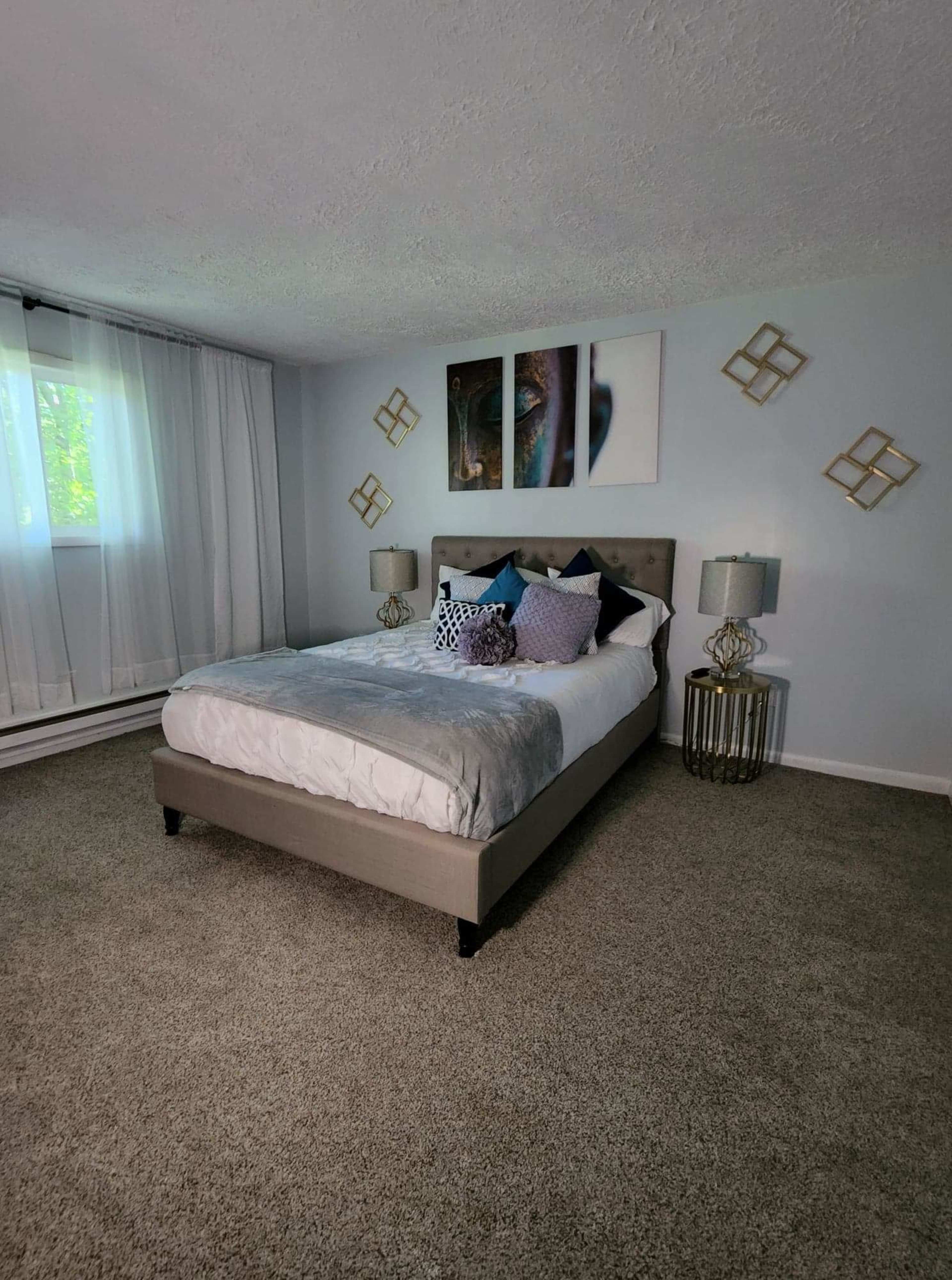 A neatly made bed with decorative pillows in a room featuring light-colored walls, lamps on bedside tables, and artwork above the bed.