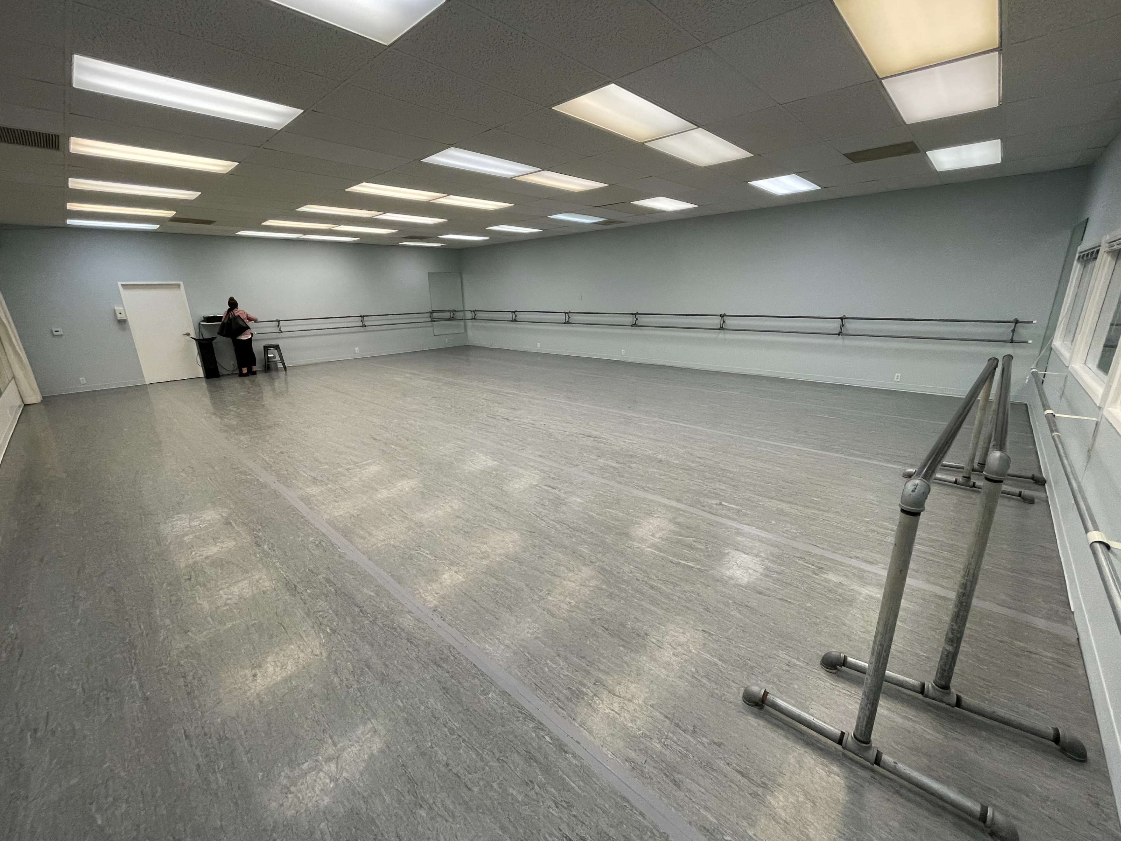 A spacious dance studio features gray flooring, mirrored walls, and a ballet bar along one side, with a person seated on a stool.