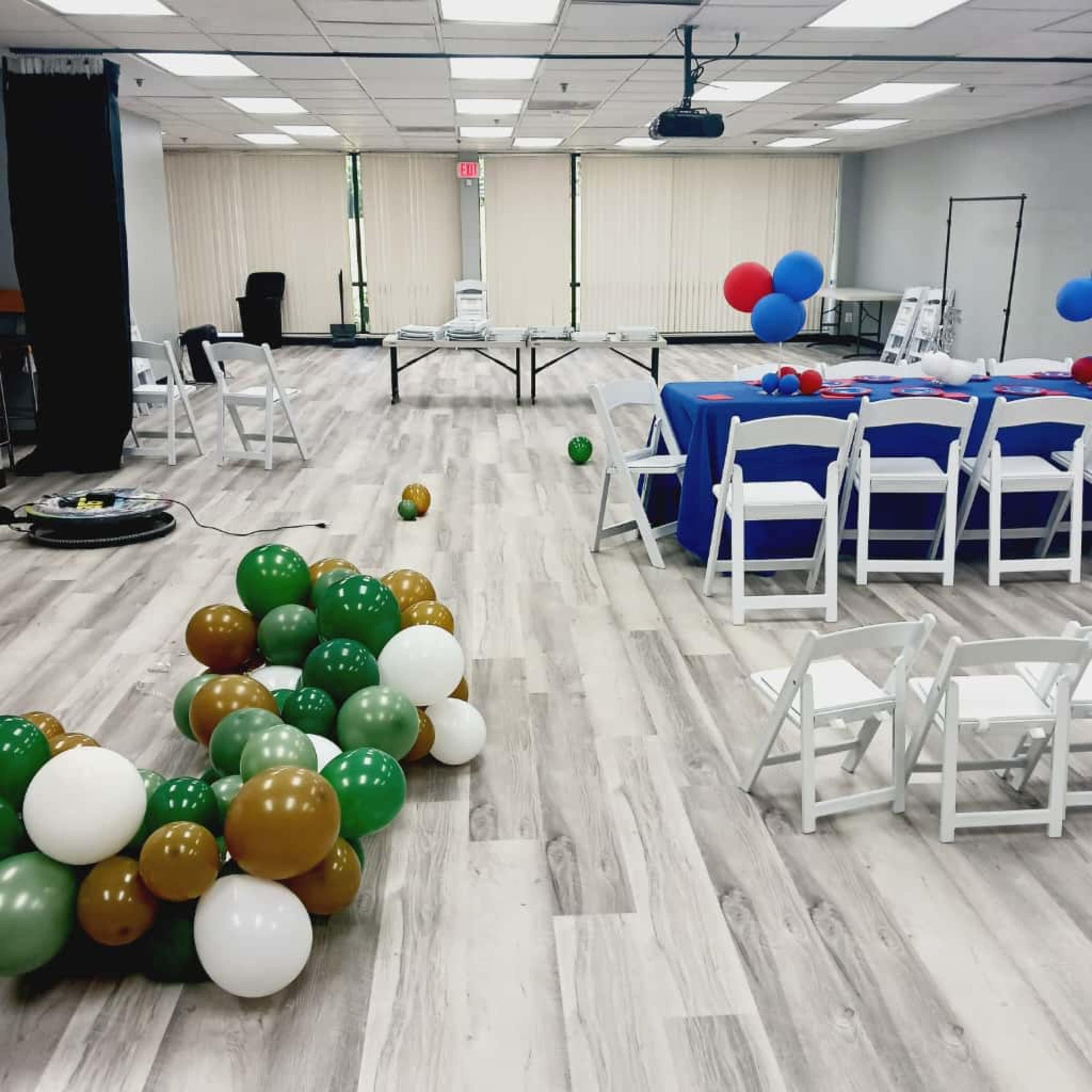 The image shows a spacious room set up for an event, featuring a table with a blue tablecloth, several white chairs, and scattered balloons in green, brown, white, and blue.
