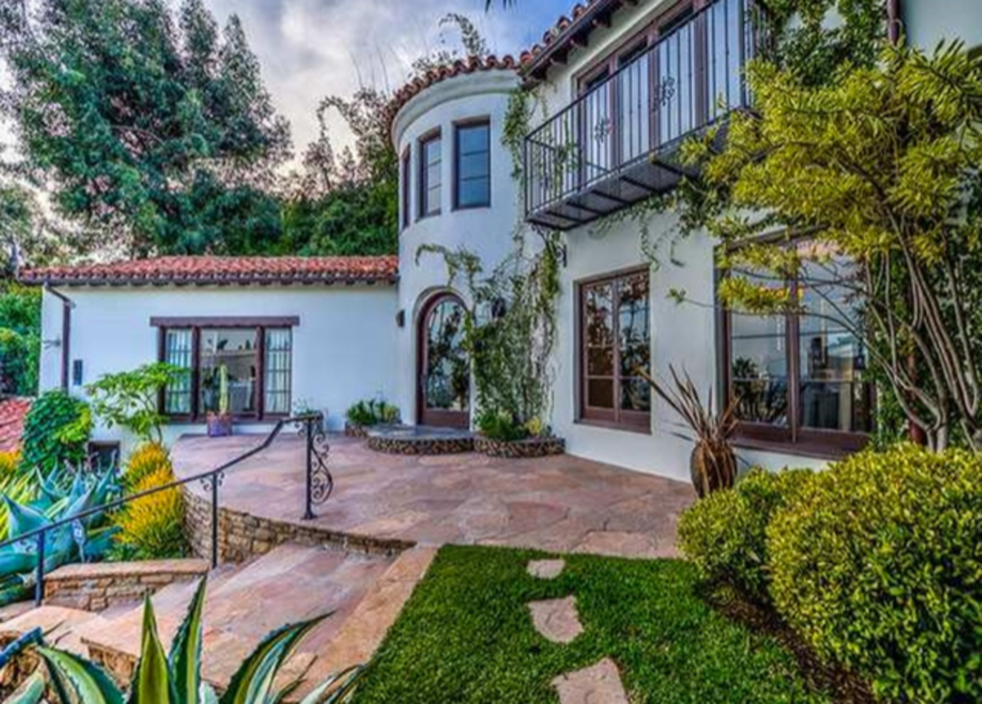 The image shows a two-story house with a stucco exterior, a tiled roof, and landscaped pathways leading to a front entrance surrounded by greenery.