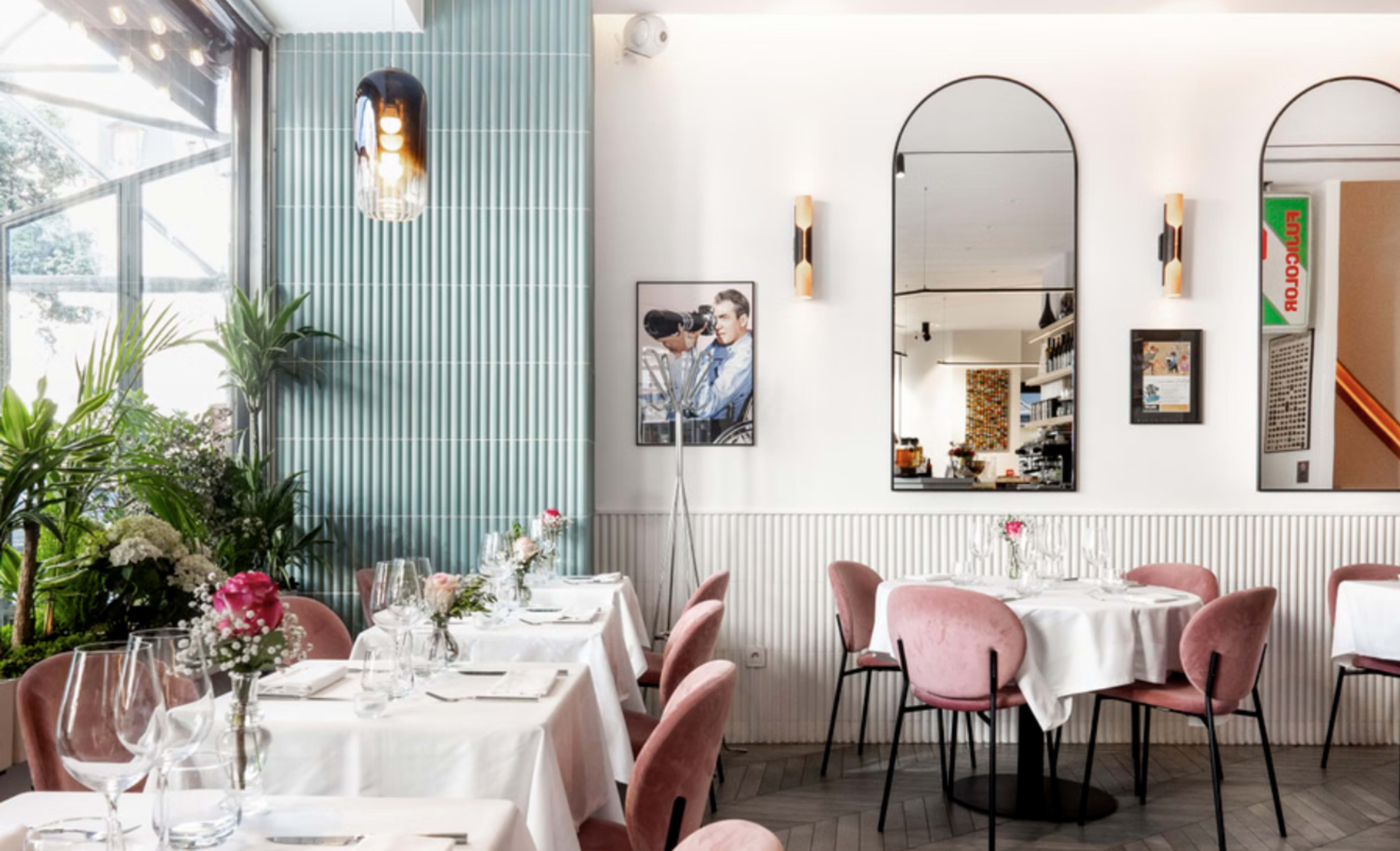 The image shows a stylish restaurant interior with pink upholstered chairs, neatly set tables, green plants, and wall-mounted mirrors.