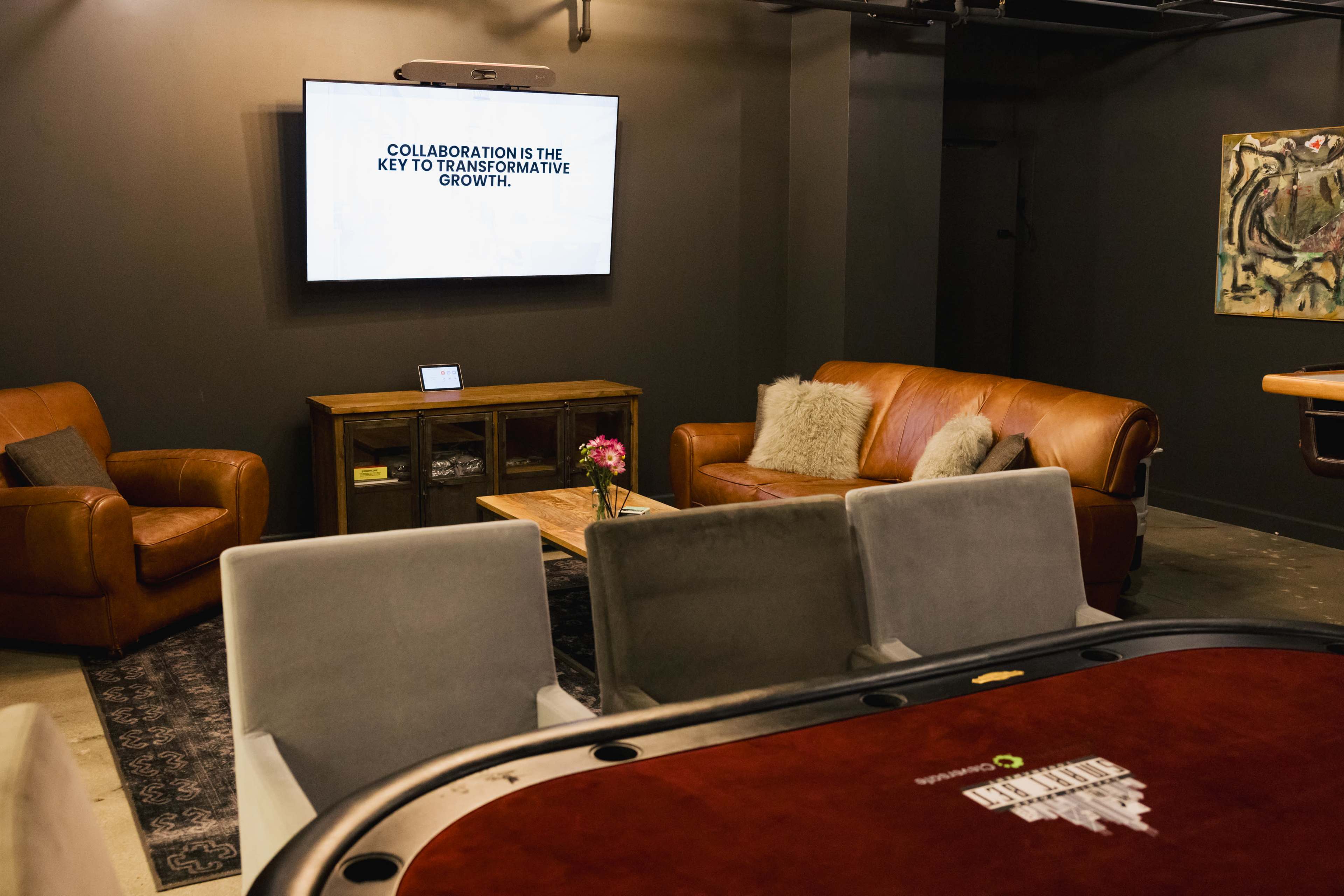 The image shows a lounge area with a large screen displaying a message about collaboration, comfortable seating, a wooden coffee table with a flower, and a poker table in the foreground.