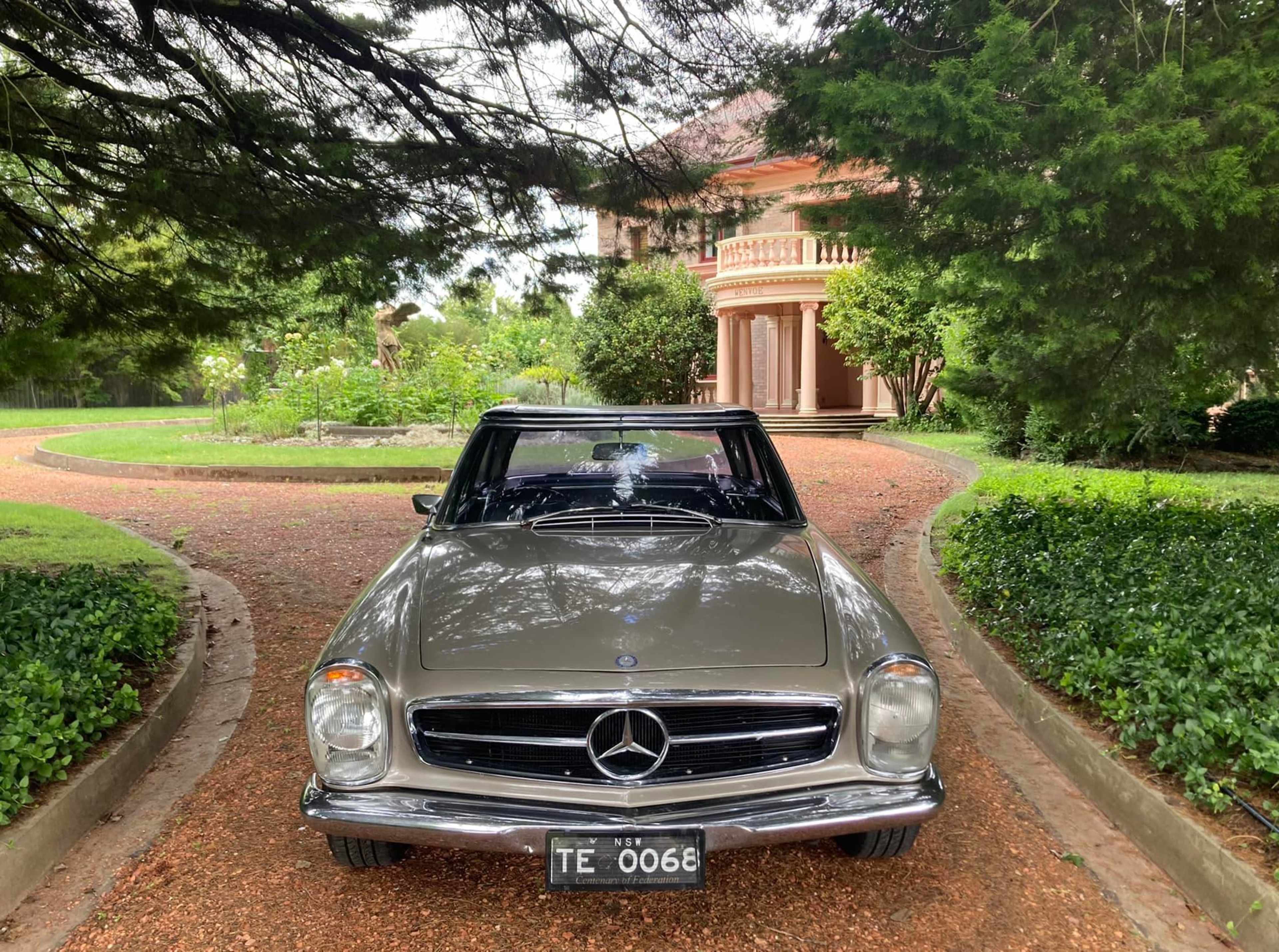 A vintage Mercedes-Benz is parked on a gravel driveway leading to a large, elegant house surrounded by lush greenery.