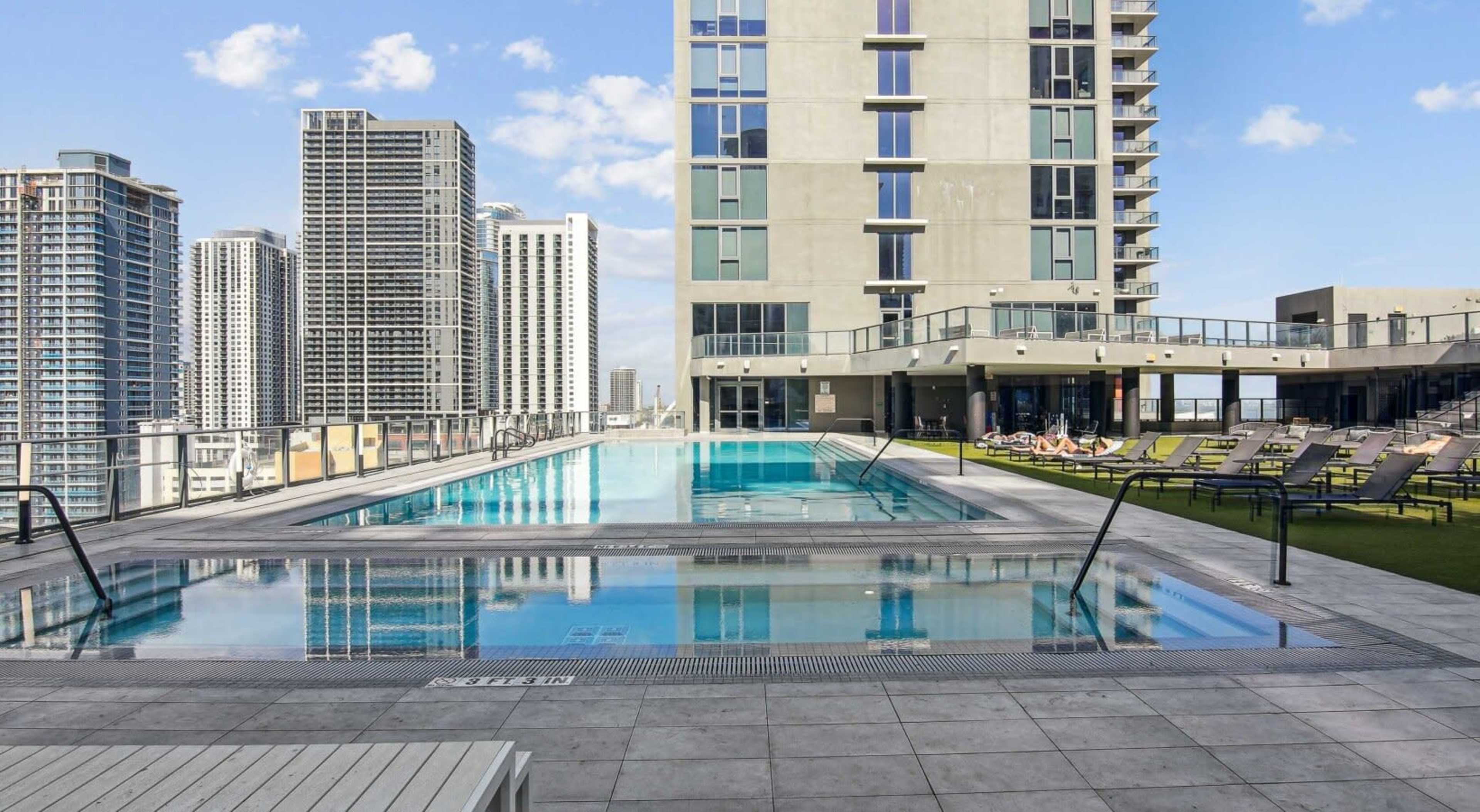 The image shows a rooftop pool area surrounded by tall buildings, with two swimming pools, lounge chairs, and a clear blue sky.