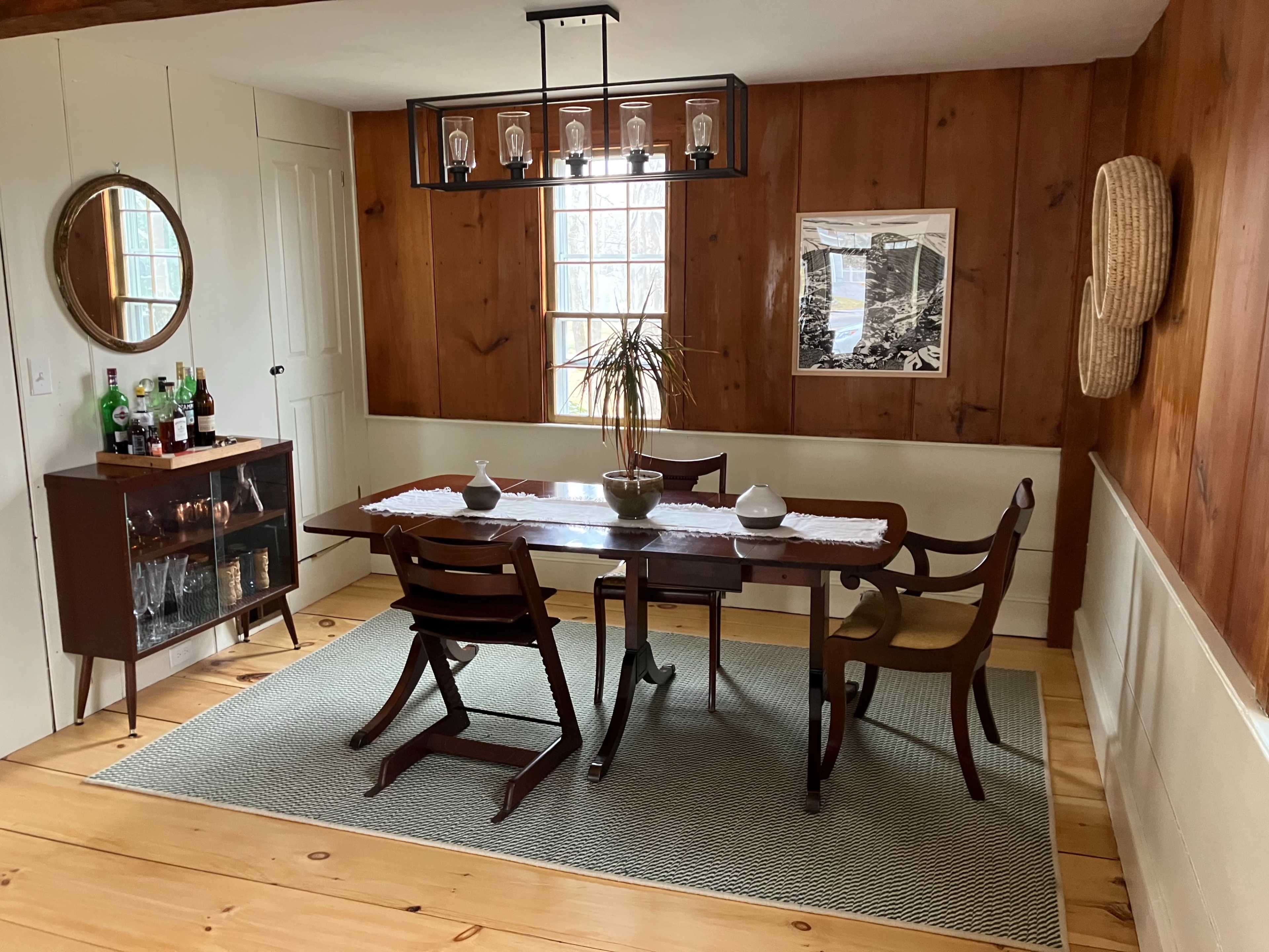 The dining room features a wooden table surrounded by chairs, a side cabinet with drinks, and large windows letting in natural light.