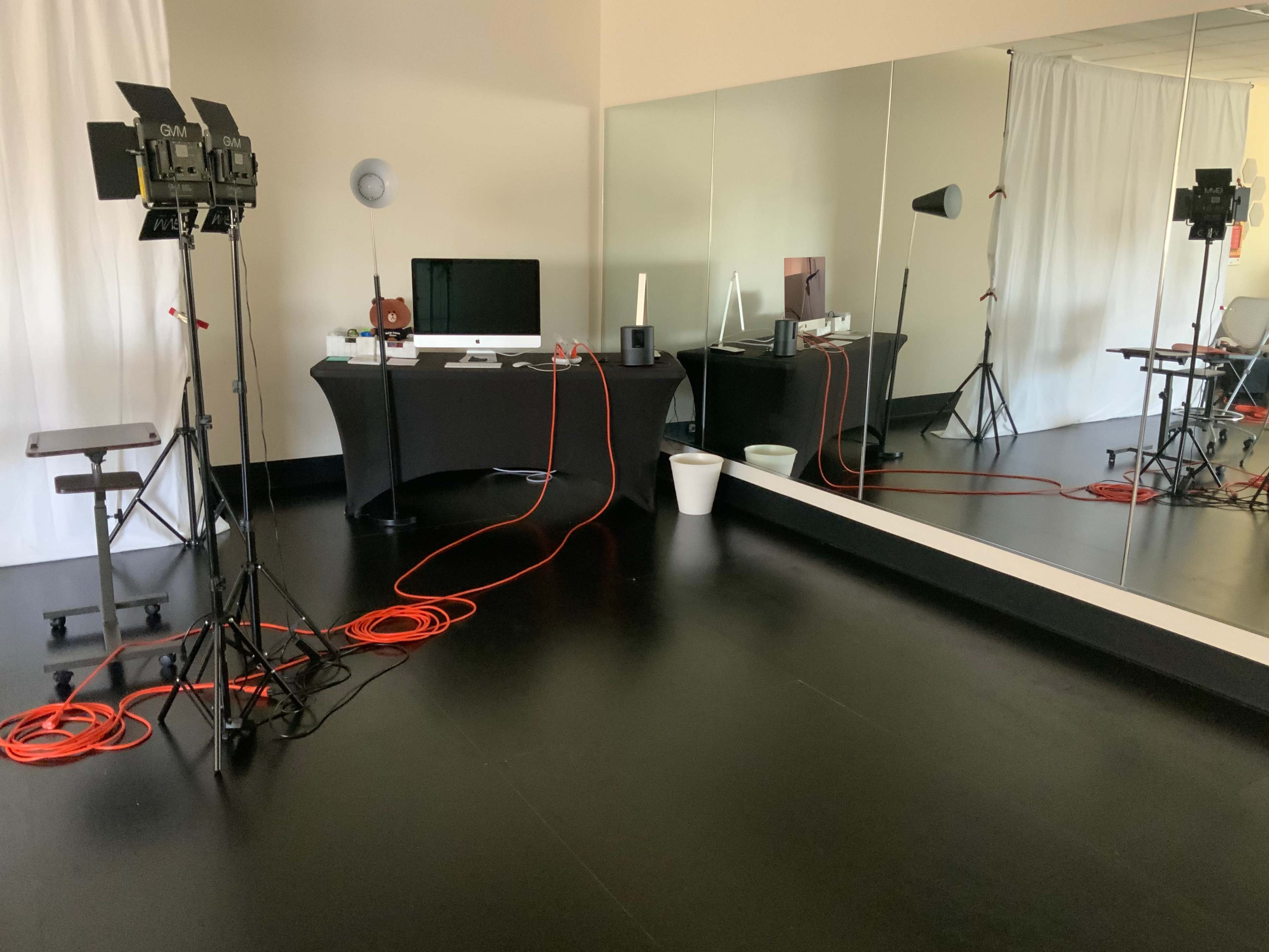A studio setting with lighting equipment, a computer on a table, and a large mirror along one wall.