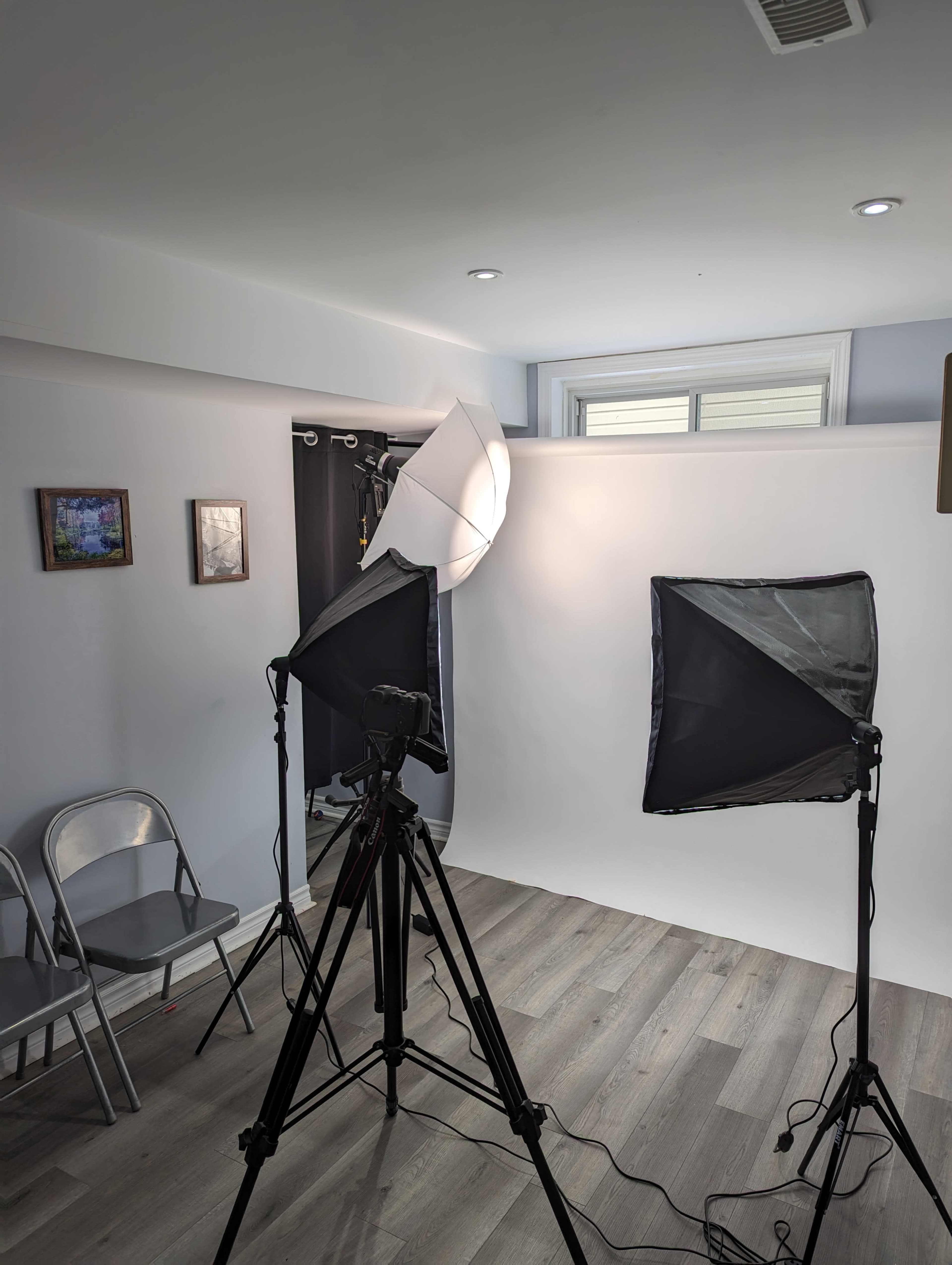 A photography studio setup with two softbox lights, a camera on a tripod, and metallic chairs nearby.