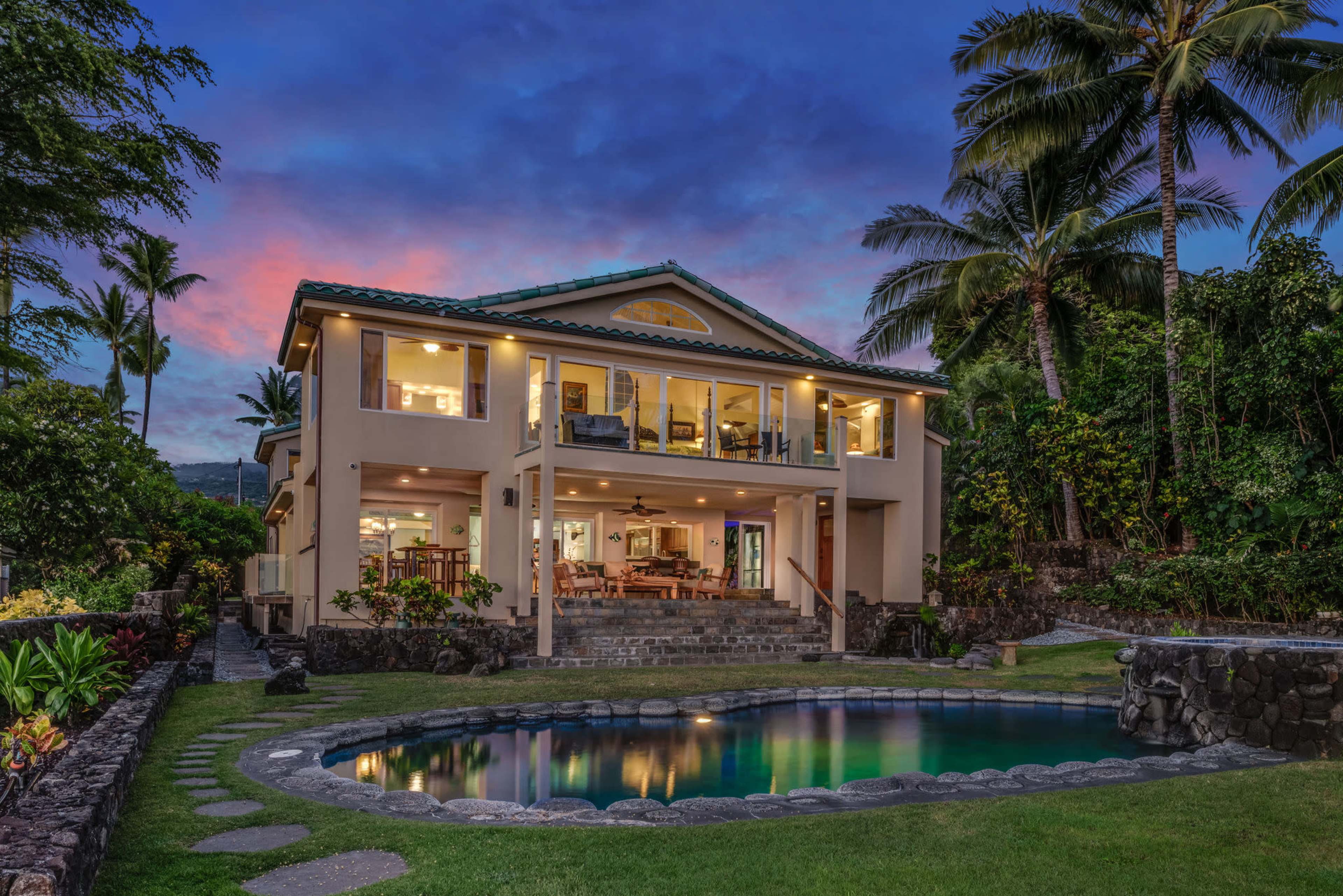 A two-story house surrounded by tropical vegetation, with a swimming pool in the foreground and a colorful sky at dusk.