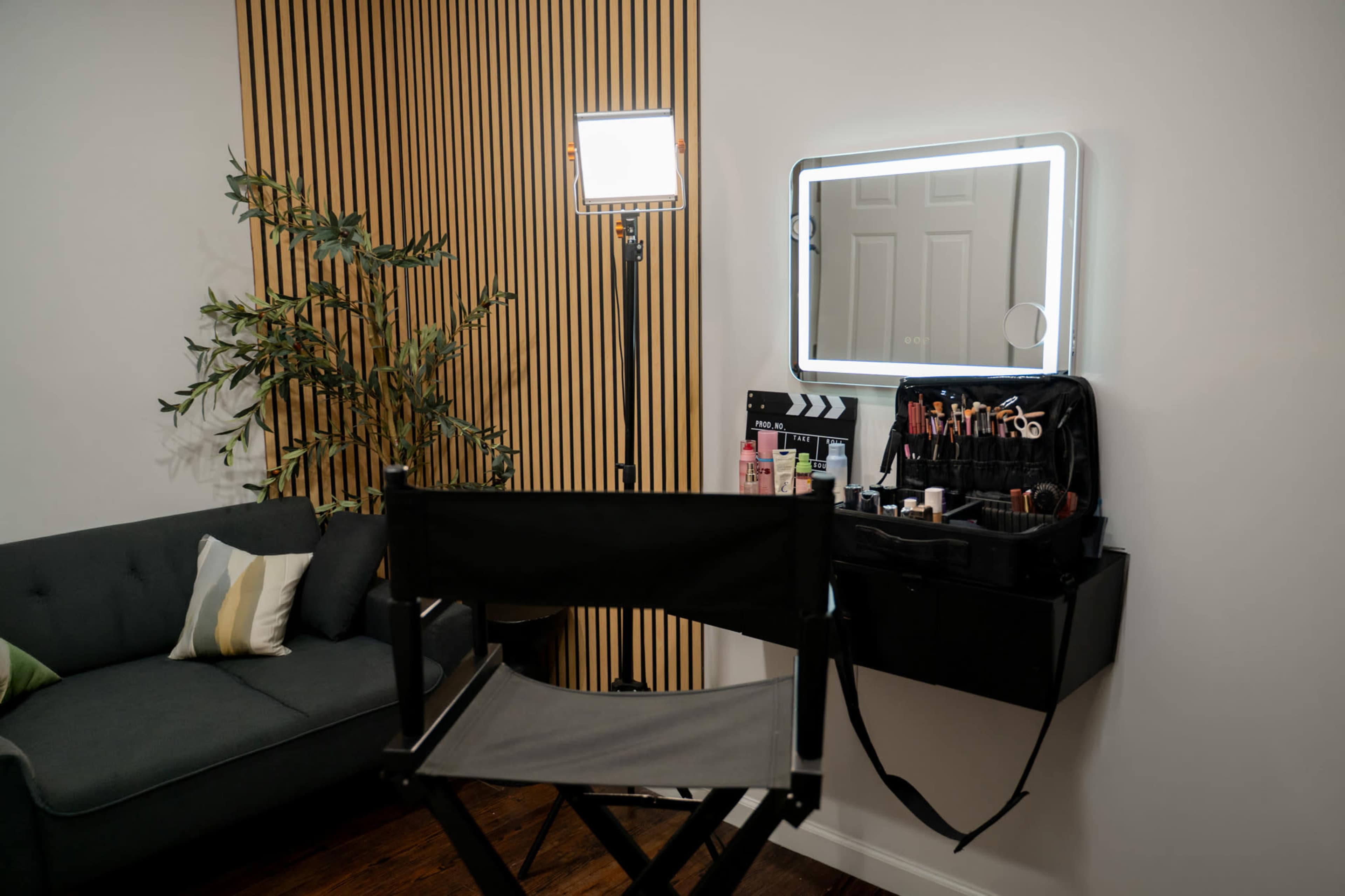 A makeup station is set up with a mirror, lighting, and organized cosmetics next to a couch and a wall with wooden paneling.