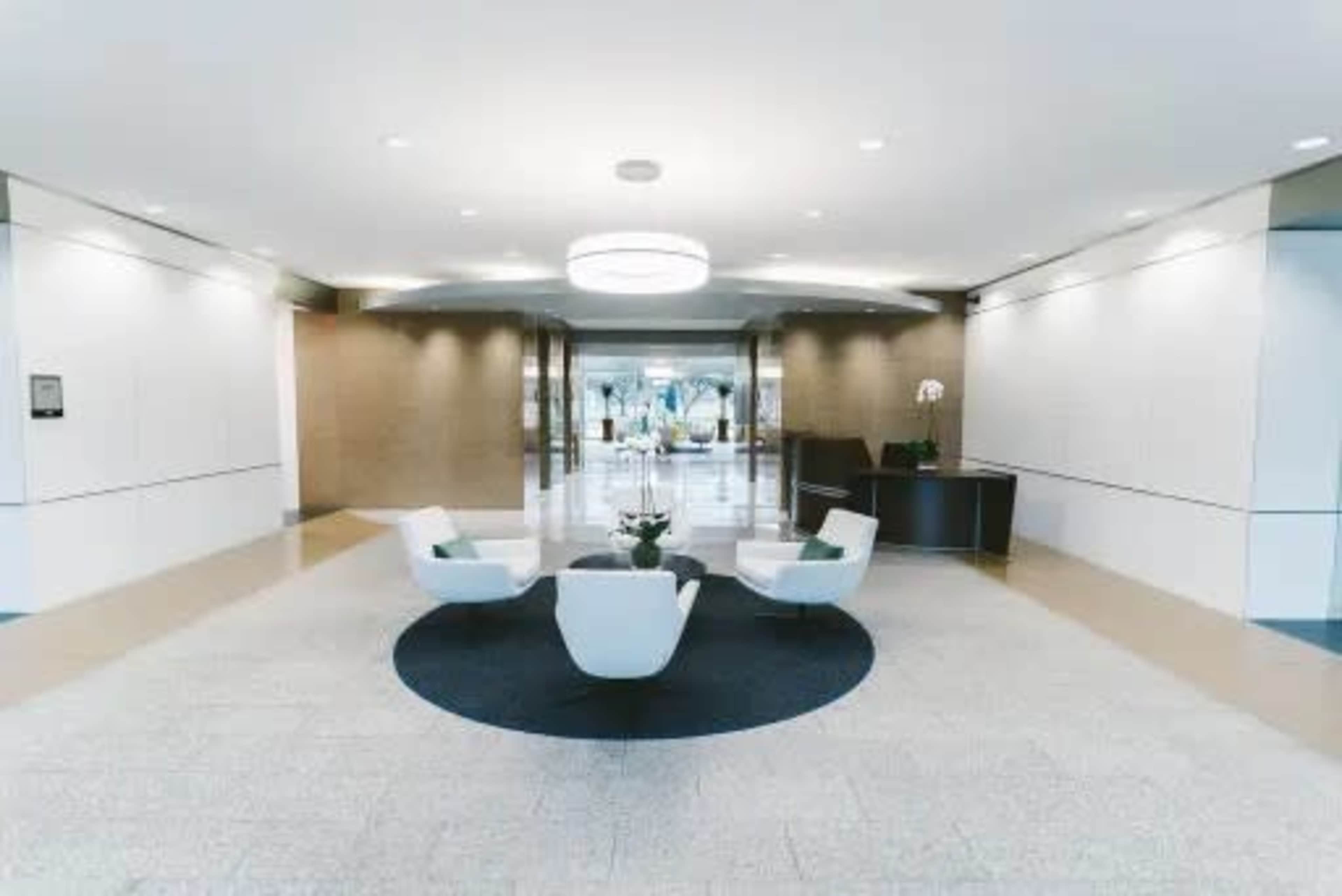 The image shows a modern reception area with three white chairs arranged around a circular black rug, set against light-colored walls and flooring.
