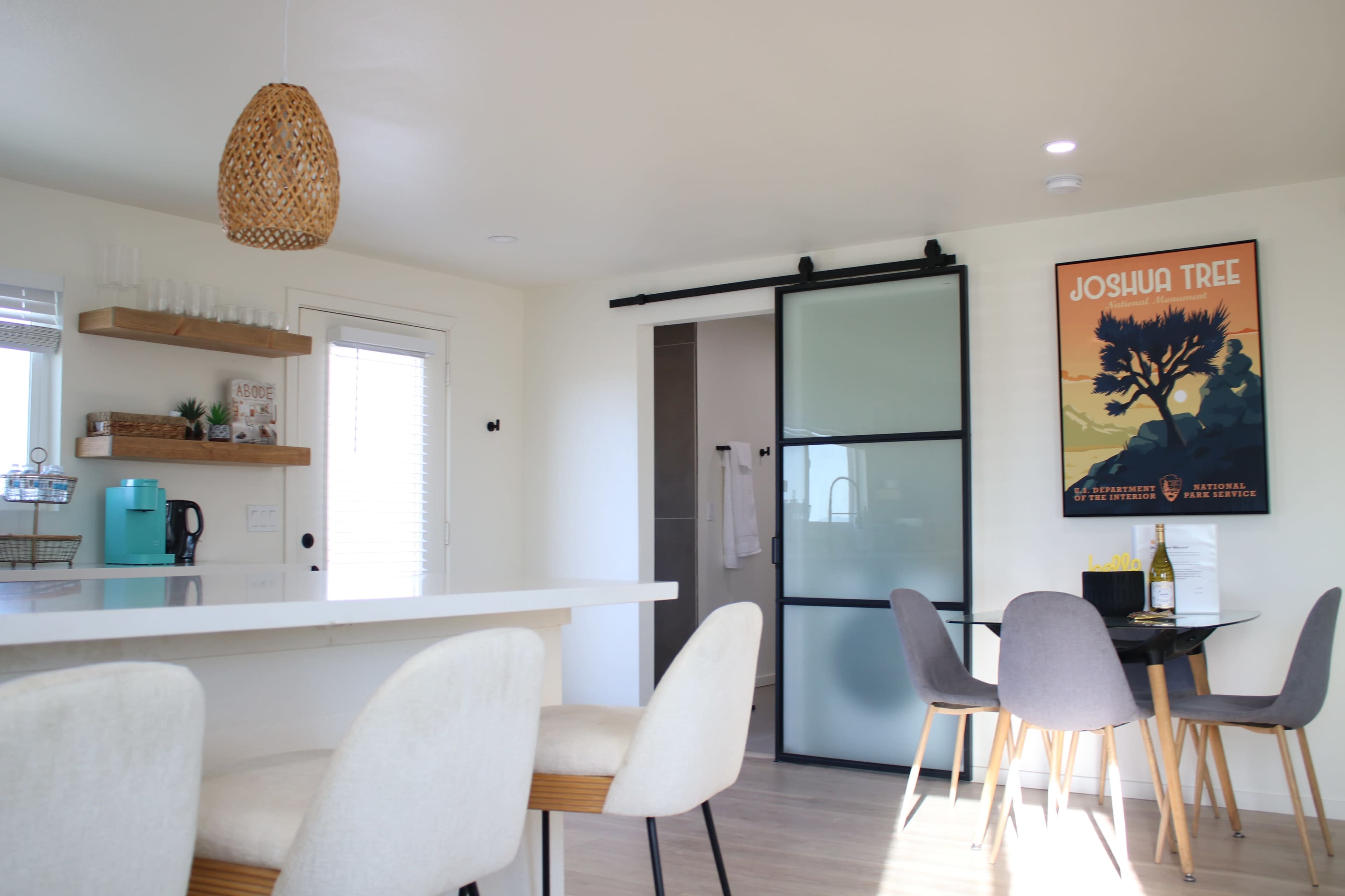 A modern open-concept kitchen and dining area features a sliding glass door, a circular dining table, and a wall-mounted Joshua Tree poster.