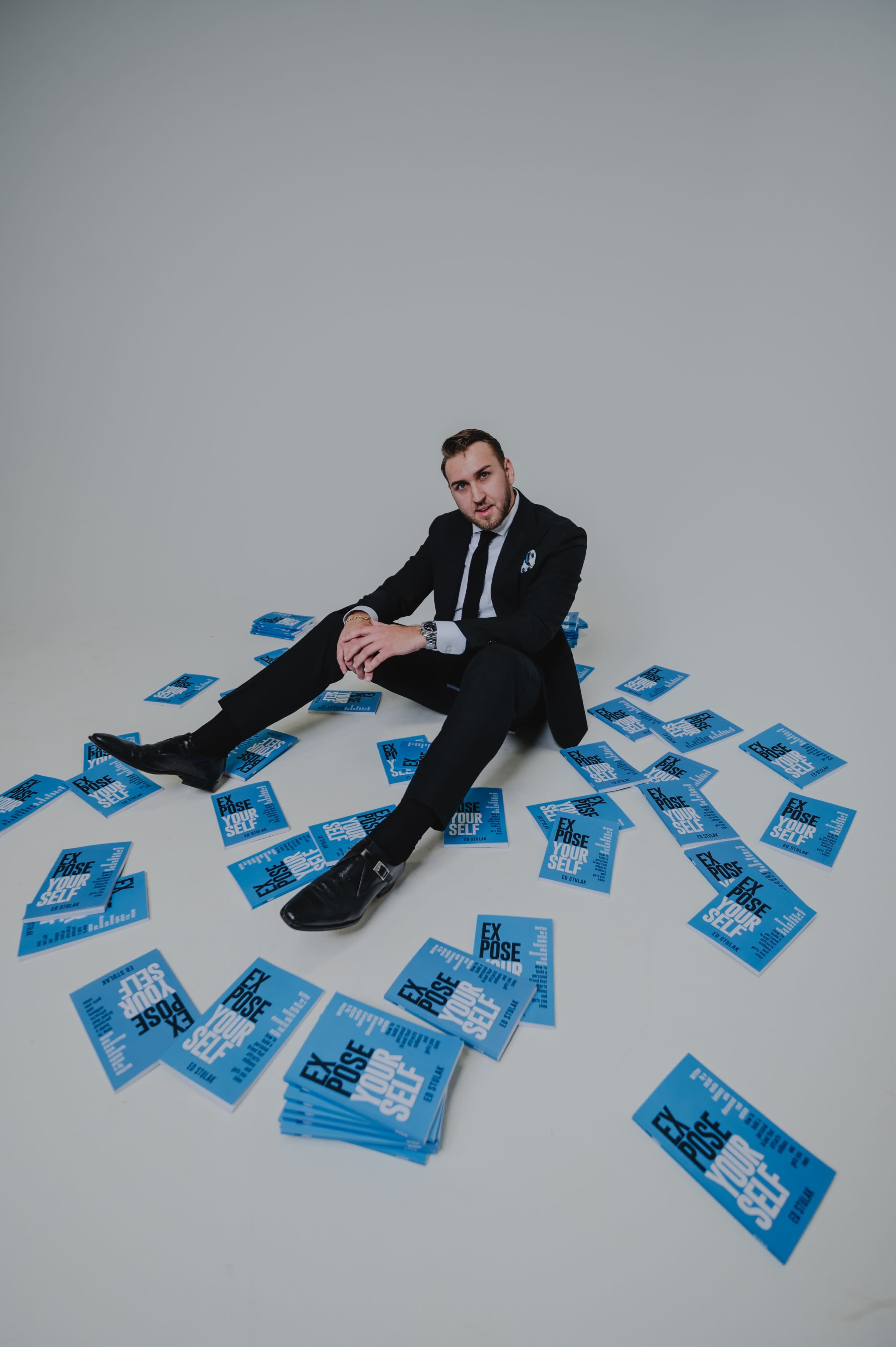 A man in a black suit sits on the floor surrounded by scattered blue books.
