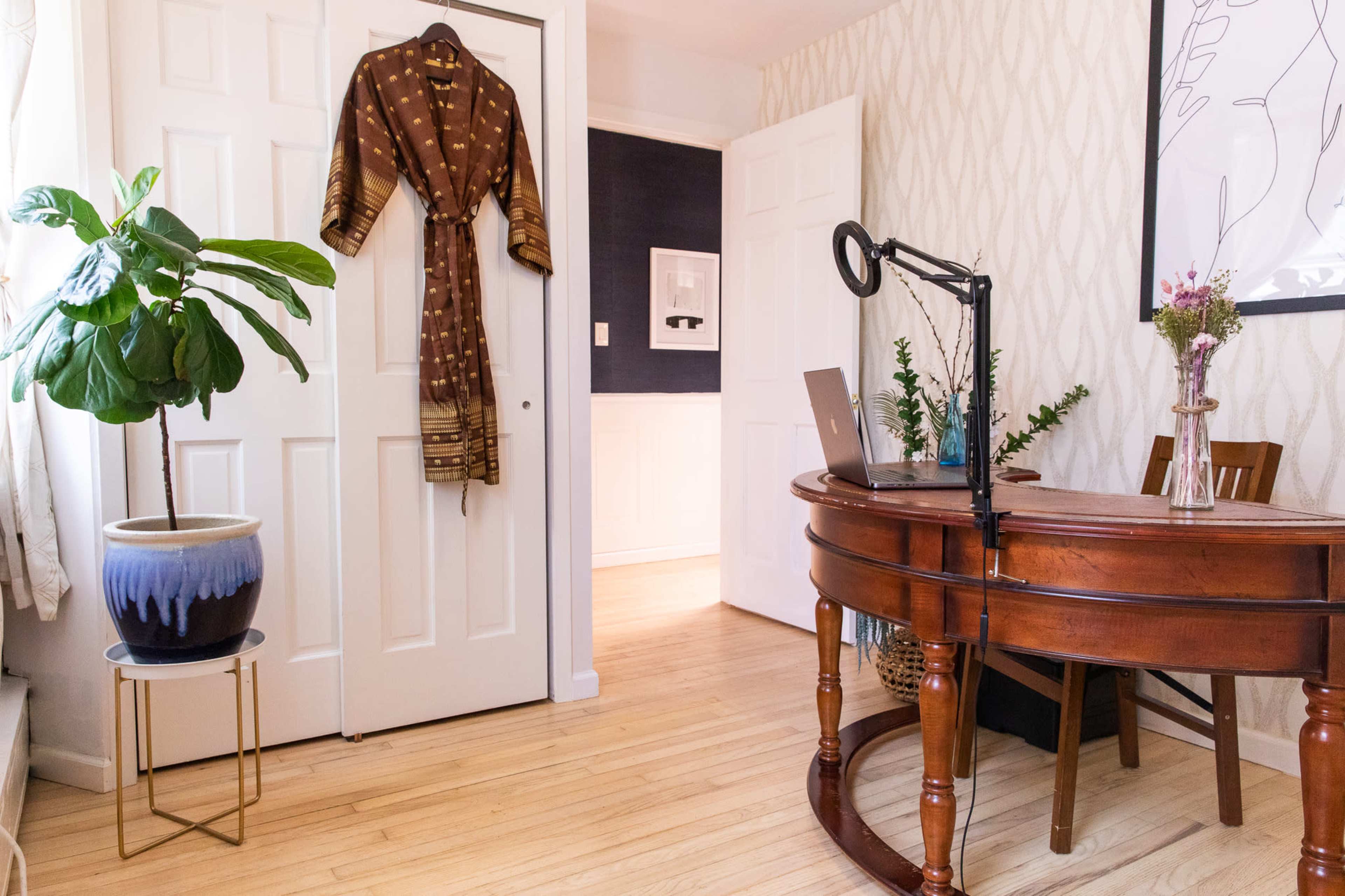 A wooden desk with a laptop is positioned in a brightly lit room featuring a plant, a decorative robe hanging on a closet door, and a chair nearby.