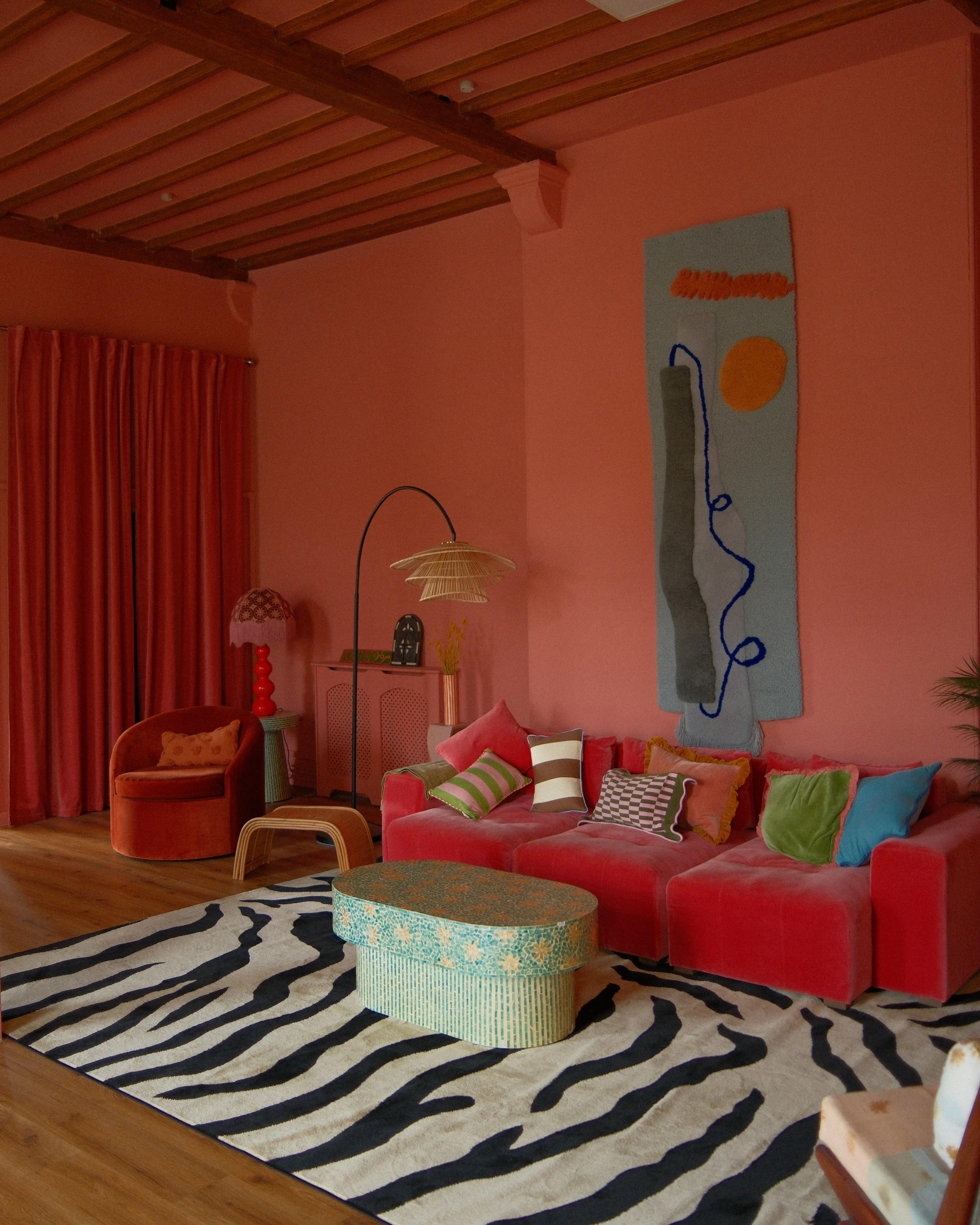 The image shows a brightly colored living room featuring a pink wall, a red sofa with various cushions, a zebra-striped rug, and decorative artwork hanging on the wall.