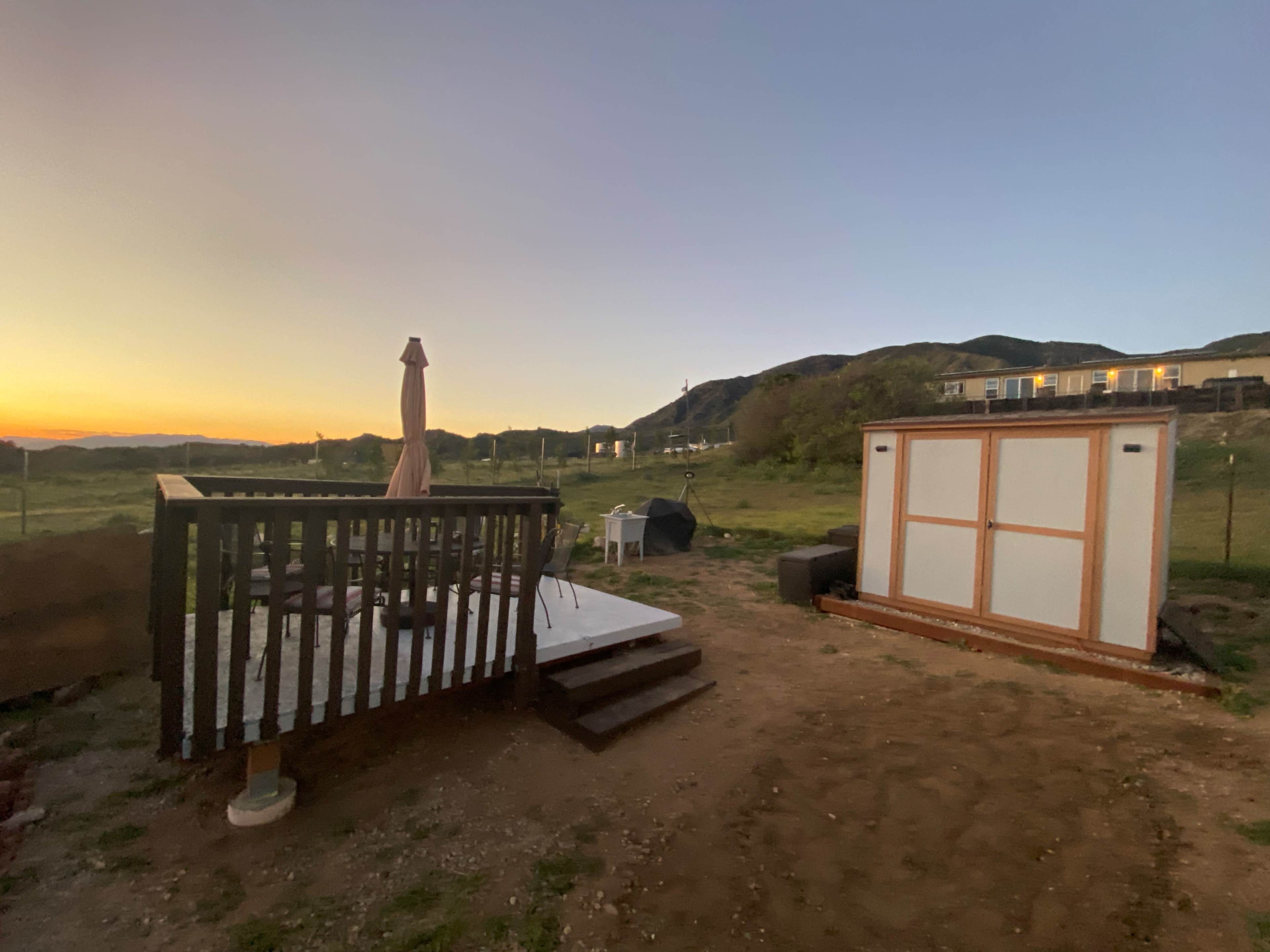 A wooden deck with a parasol and a storage shed is situated in an open area at dusk, with hills and buildings in the background.