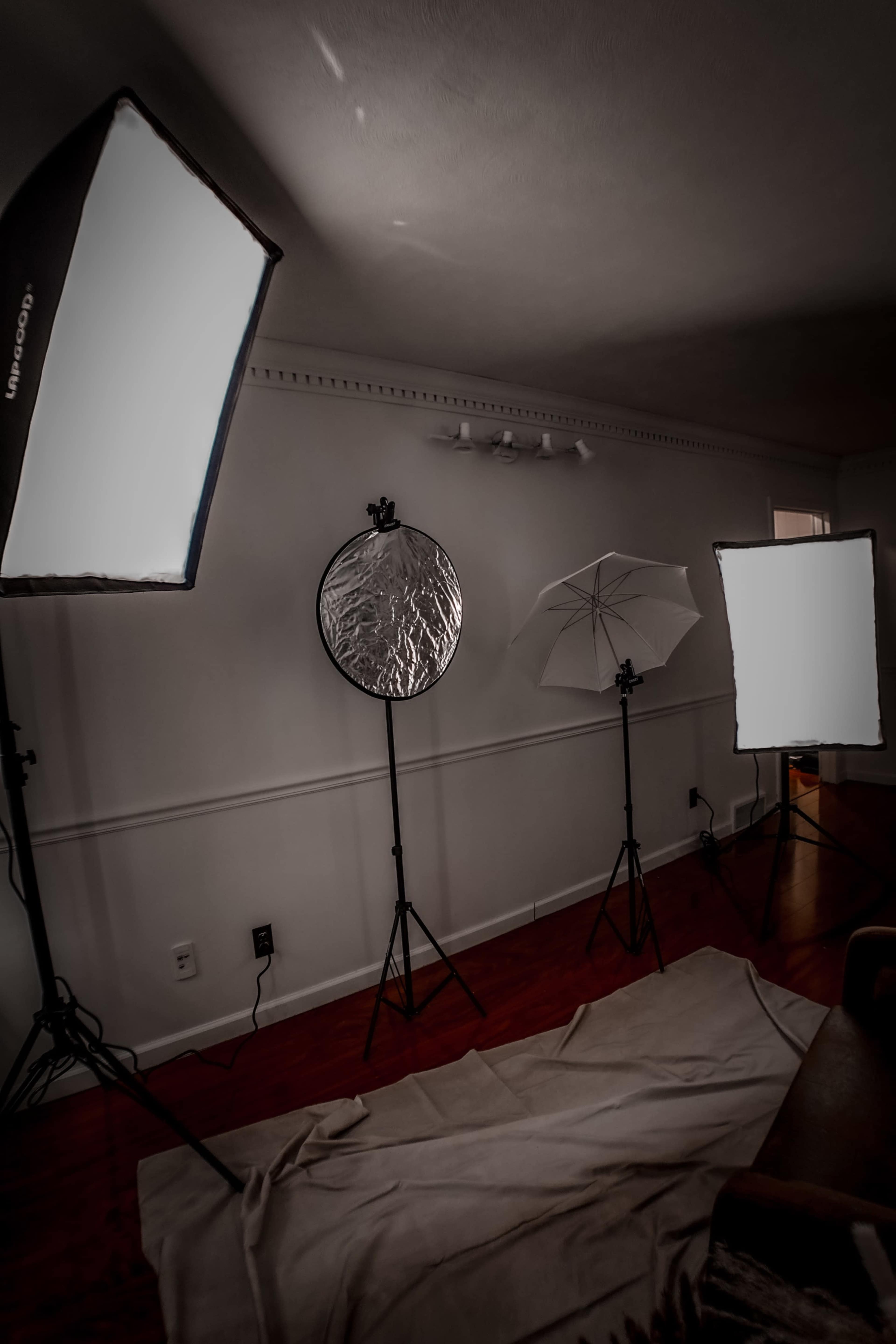 The image shows a dimly lit room with three lighting setups and a reflector arranged on light stands, along with a white cloth laid on the floor.