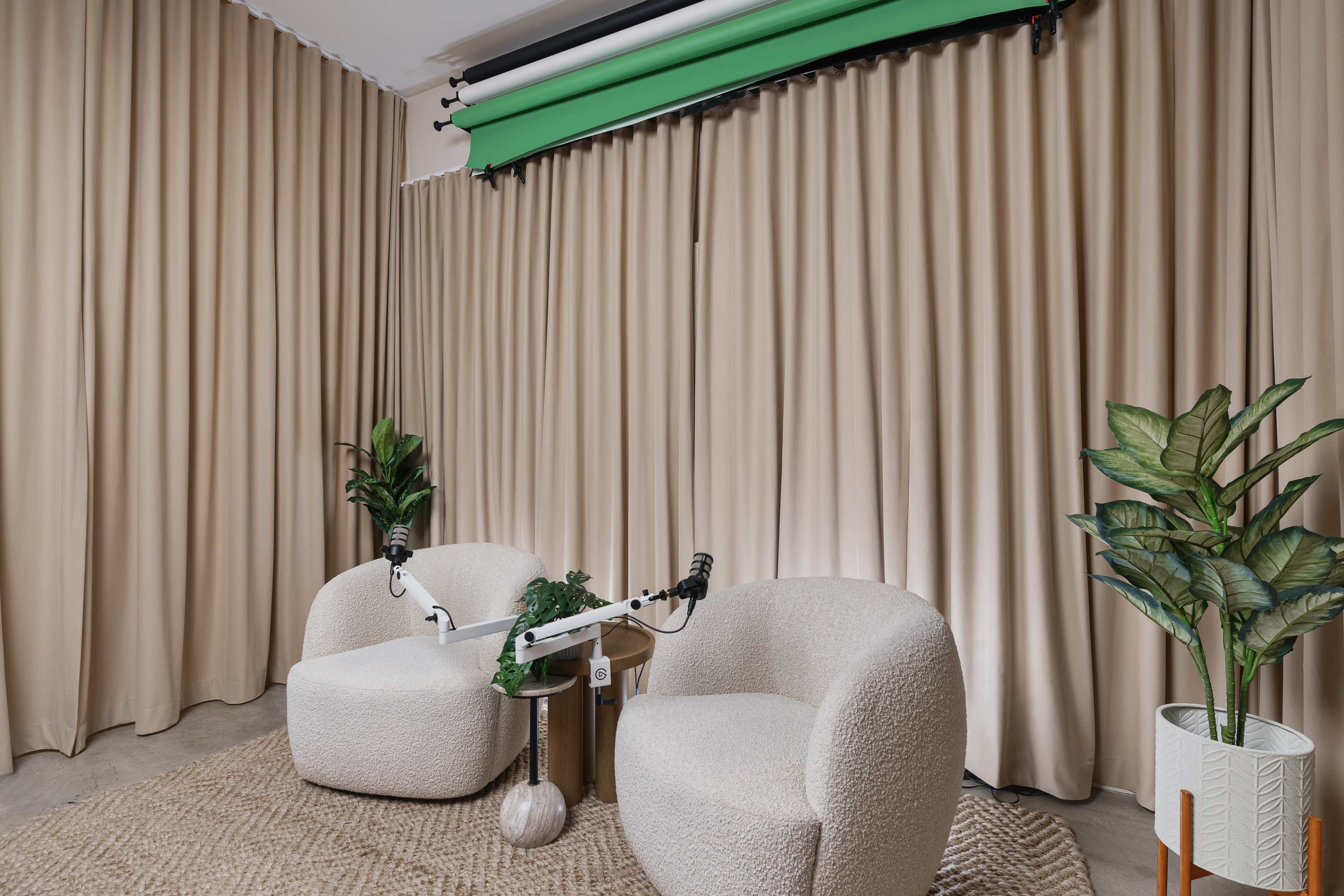 A modern recording space with two white upholstered chairs, a green backdrop, and potted plants against beige curtains.