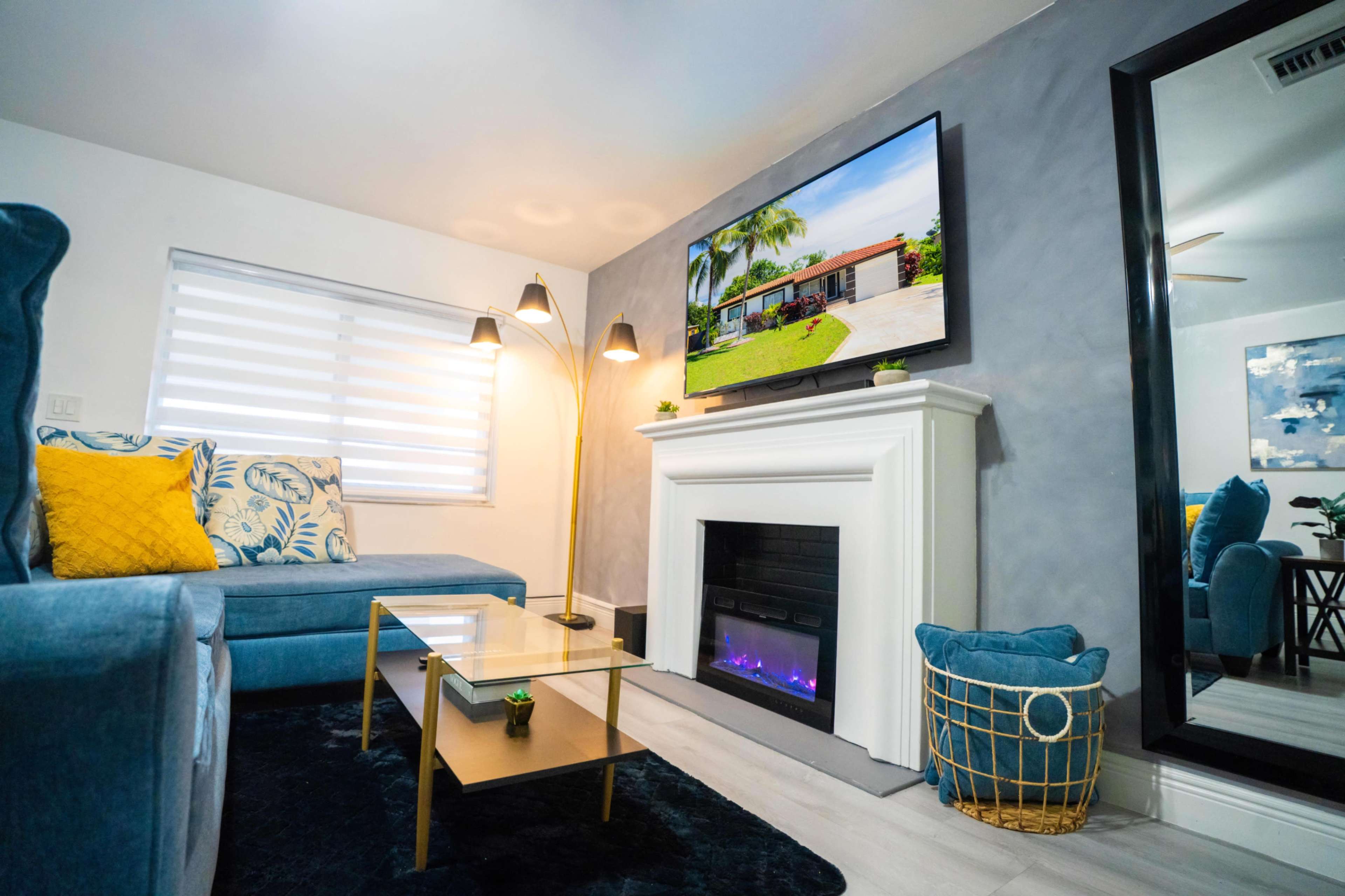 The image shows a modern living room with a blue sectional sofa, a glass coffee table, a decorative fireplace, and a wall-mounted television.