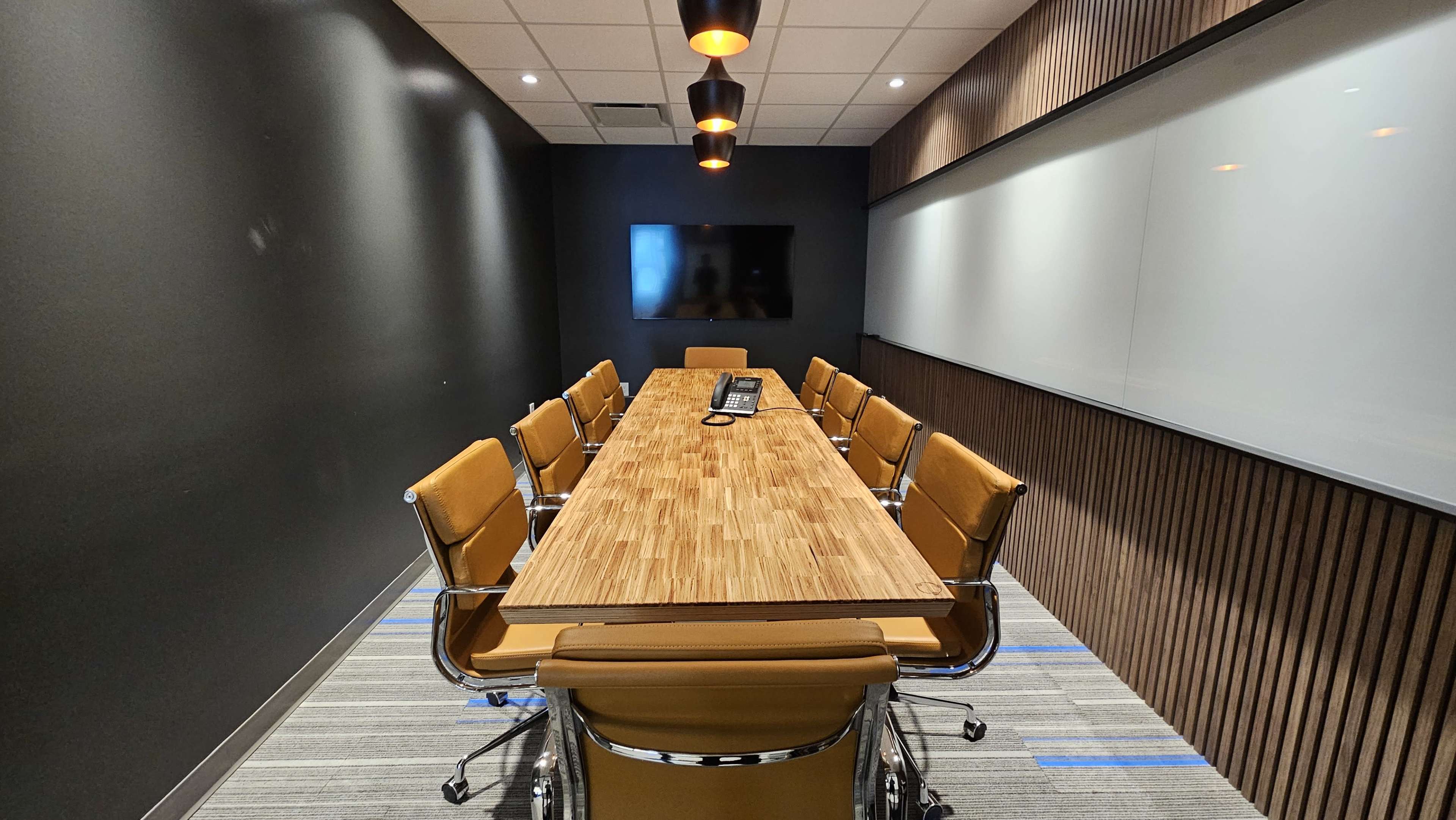 A long wooden conference table with orange chairs is set in a modern meeting room featuring a dark wall and a large screen.